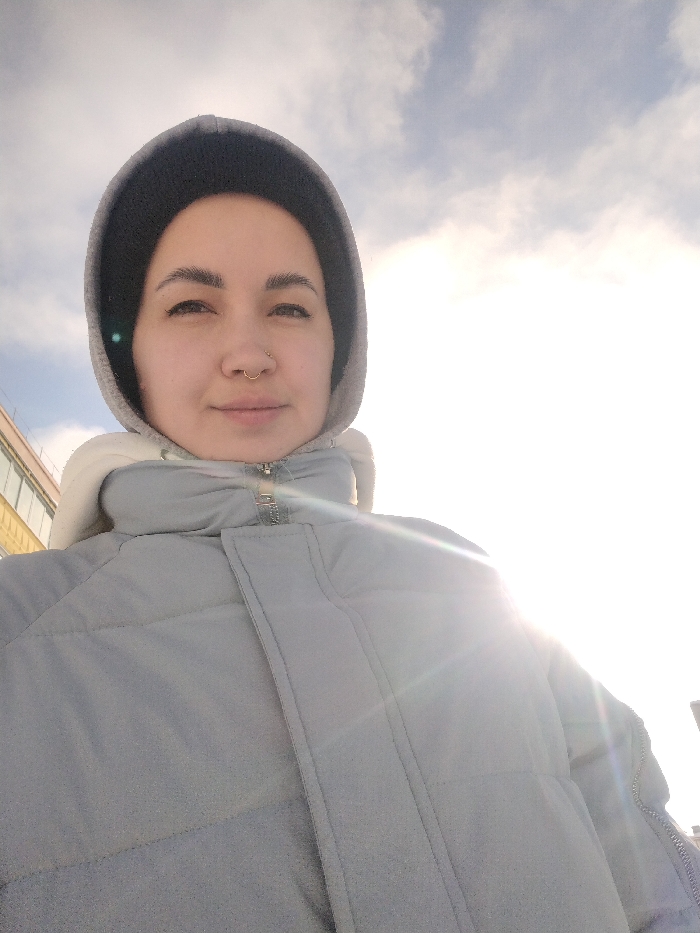 The image is a bright, upward-angled outdoor selfie or close-up portrait featuring a person, likely female. She is positioned towards the left side of the frame, looking directly at the camera with a neutral and calm expression.
She has dark, well-defined eyebrows and appears to have a septum piercing. Her hair is mostly covered by a dark grey or black hood, which is then covered by a lighter grey hood, suggesting multiple layers of clothing. She is wearing a thick, light grey or light blue puffer jacket with a prominent zipper and visible stitching that creates quilted sections.
The background is dominated by a bright sky, with scattered white clouds visible mostly in the upper left portion. A very strong light source, presumably the sun, is positioned in the lower right of the frame, causing significant lens flare and creating a large area of bright overexposure on the right side of the image. This backlighting casts a glow around the person's hood and shoulder. On the far left, a blurred portion of a building with visible windows can be seen in the distance.
The bright sunlight indicates it is daytime, and the heavy winter jacket suggests cold weather. There is no visible text in the image.