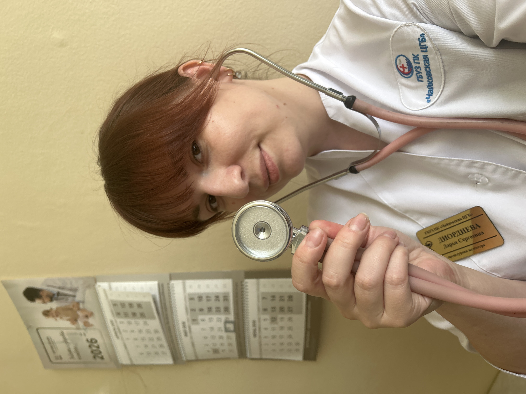A person wearing a white medical coat with a stethoscope around their neck is holding the stethoscope's head in their hand. A name tag is on the coat. The name tag contains the phrase "ДИОРДИЕВА Дарья Сергеевна,". The coat also has the phrase "ГБУЗ ПК «Чайковская ЦГБ»," on it. There is a calendar on the wall showing the year 2026. The organization name "ГБУЗ ПК «Чайковская ЦГБ»" is associated with the city of Чайковский, Russia.