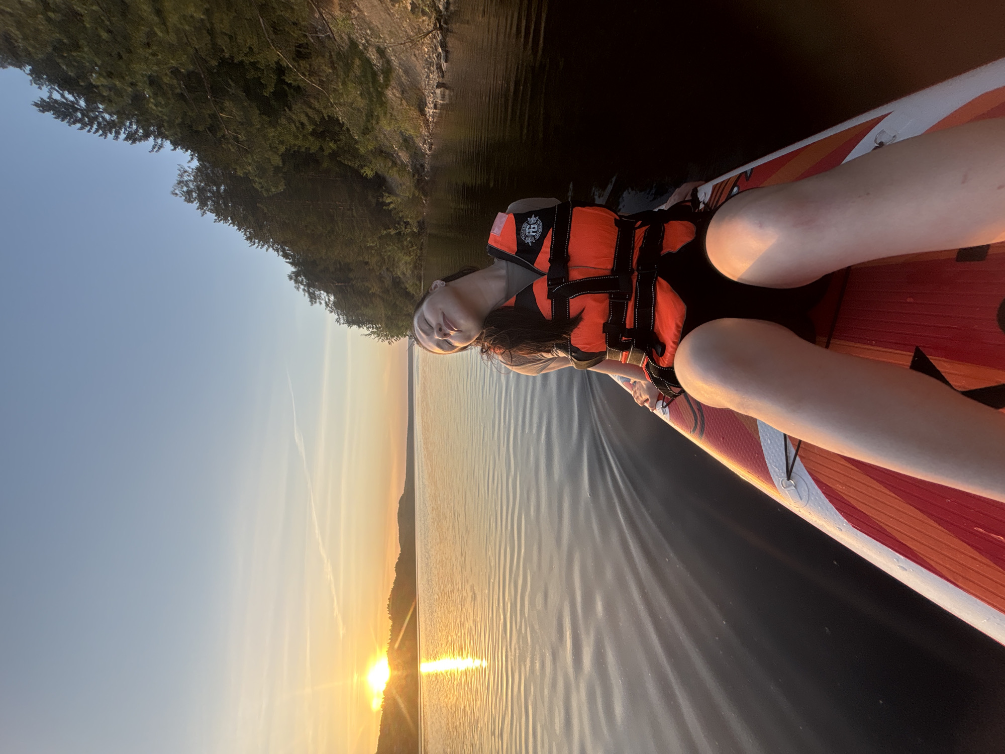 The image shows a person wearing a life jacket lying on a paddleboard in a body of water at sunset. The sun is low on the horizon, reflecting in the water. Land with trees is visible in the background. The location in the city of Khokhlovka, Russia, cannot be determined from this image.