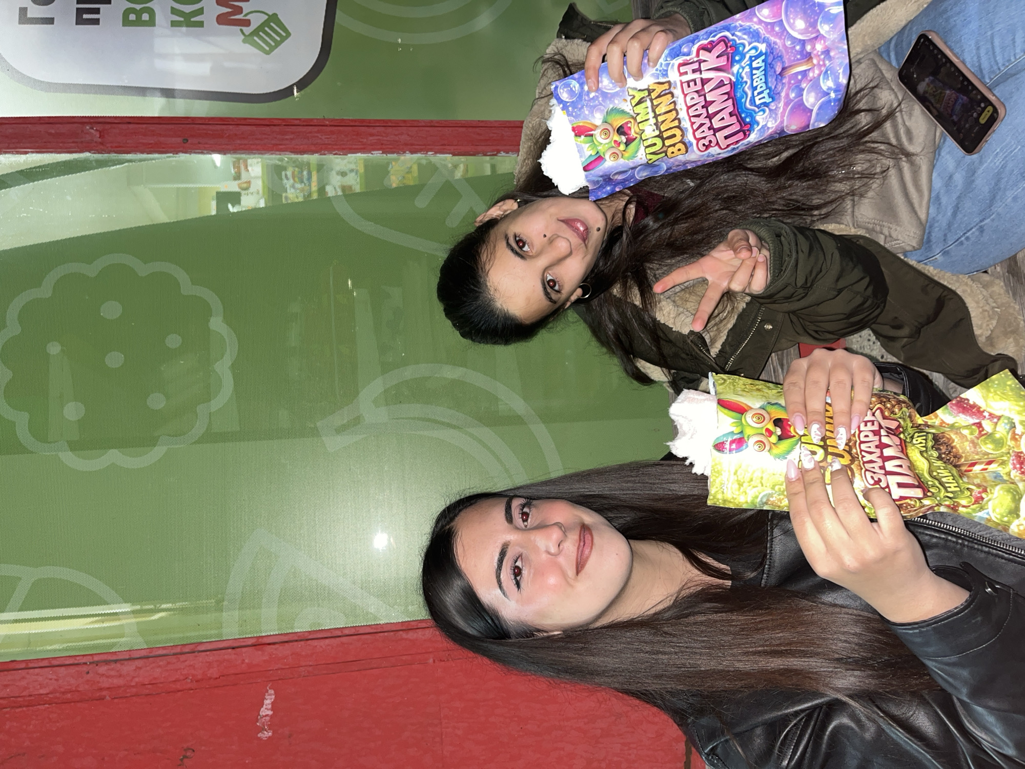 Two young women hold bags of "Yummy Bunny Zaxapeh Pamuk" candy floss in front of a window display with green, patterned graphics and a red frame. One woman is wearing a black leather jacket, and the other is wearing a beige jacket with a fur collar and blue jeans. The second woman is also making a peace sign with her hand. A phone is visible near her hand. The location is likely Sungurlare, Bulgaria, given the Cyrillic text on the candy floss packaging.