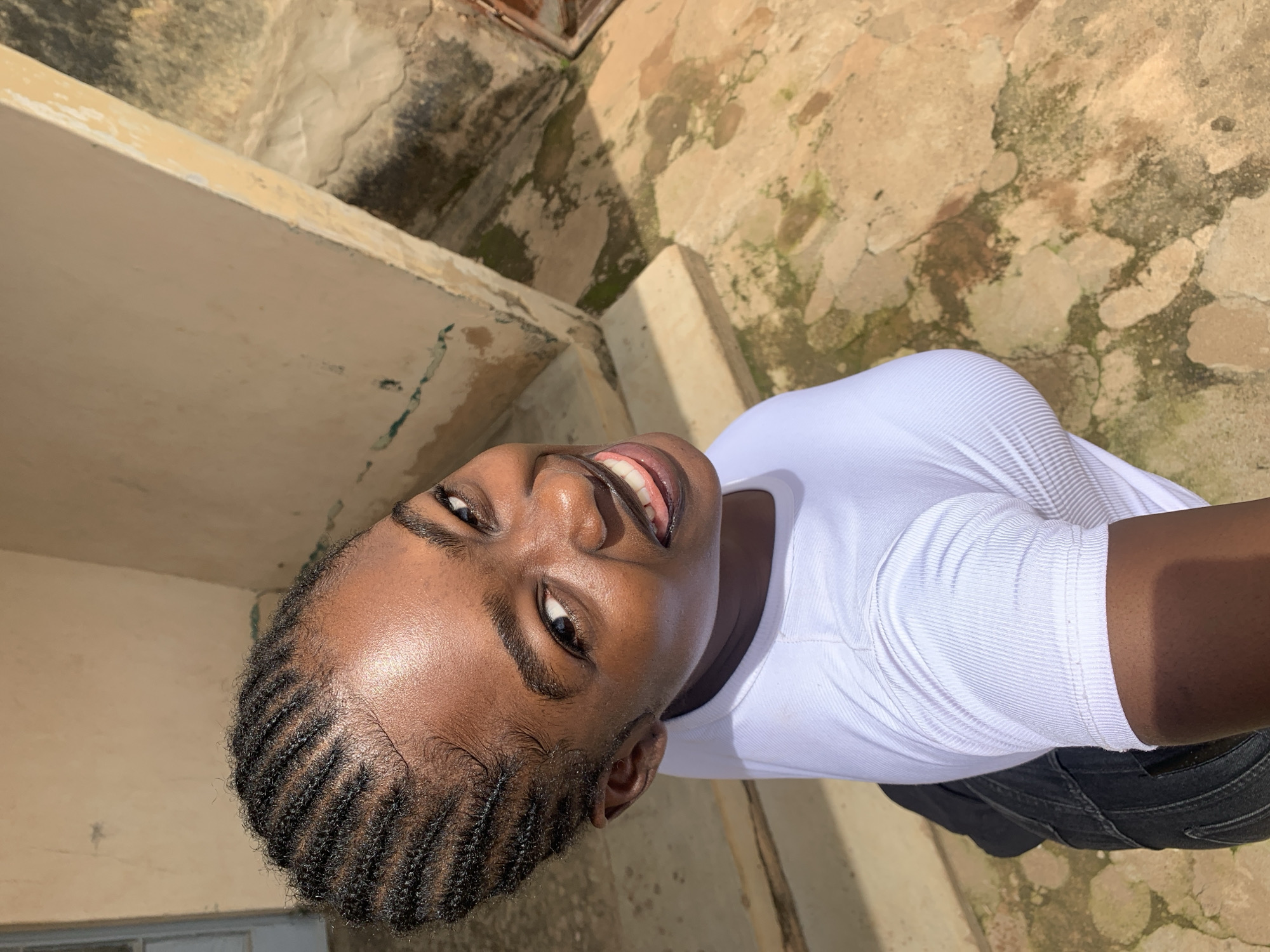 A young Black woman with braided hair smiles at the camera in an outdoor setting in Rafin Par, Nigeria. She is wearing a white ribbed t-shirt and dark pants. The background consists of a textured wall, stairs, and a stone-paved ground with patches of moss and dirt. The lighting suggests it is daytime, with sunlight casting shadows.