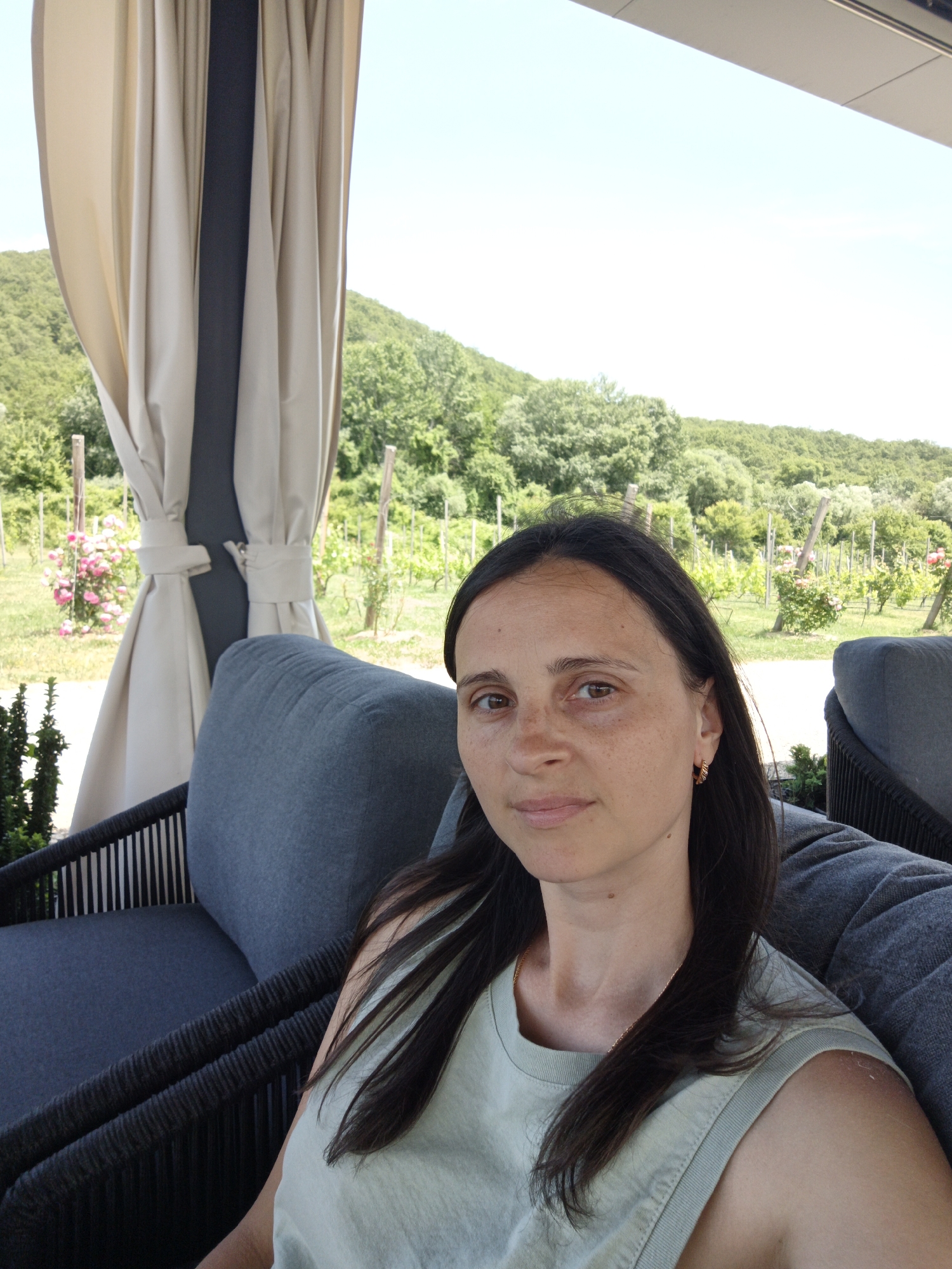 A medium shot photo shows a woman sitting on a dark gray cushioned outdoor chair. She is looking directly at the camera. Her dark brown hair is long and falls over her shoulders and chest. She is wearing a light green sleeveless top. Behind her is a vineyard with rows of grapevines and some rose bushes in bloom. In the background, green hills are covered with trees. A beige awning with gray draping is visible to the left of the woman. The sky is clear and bright.