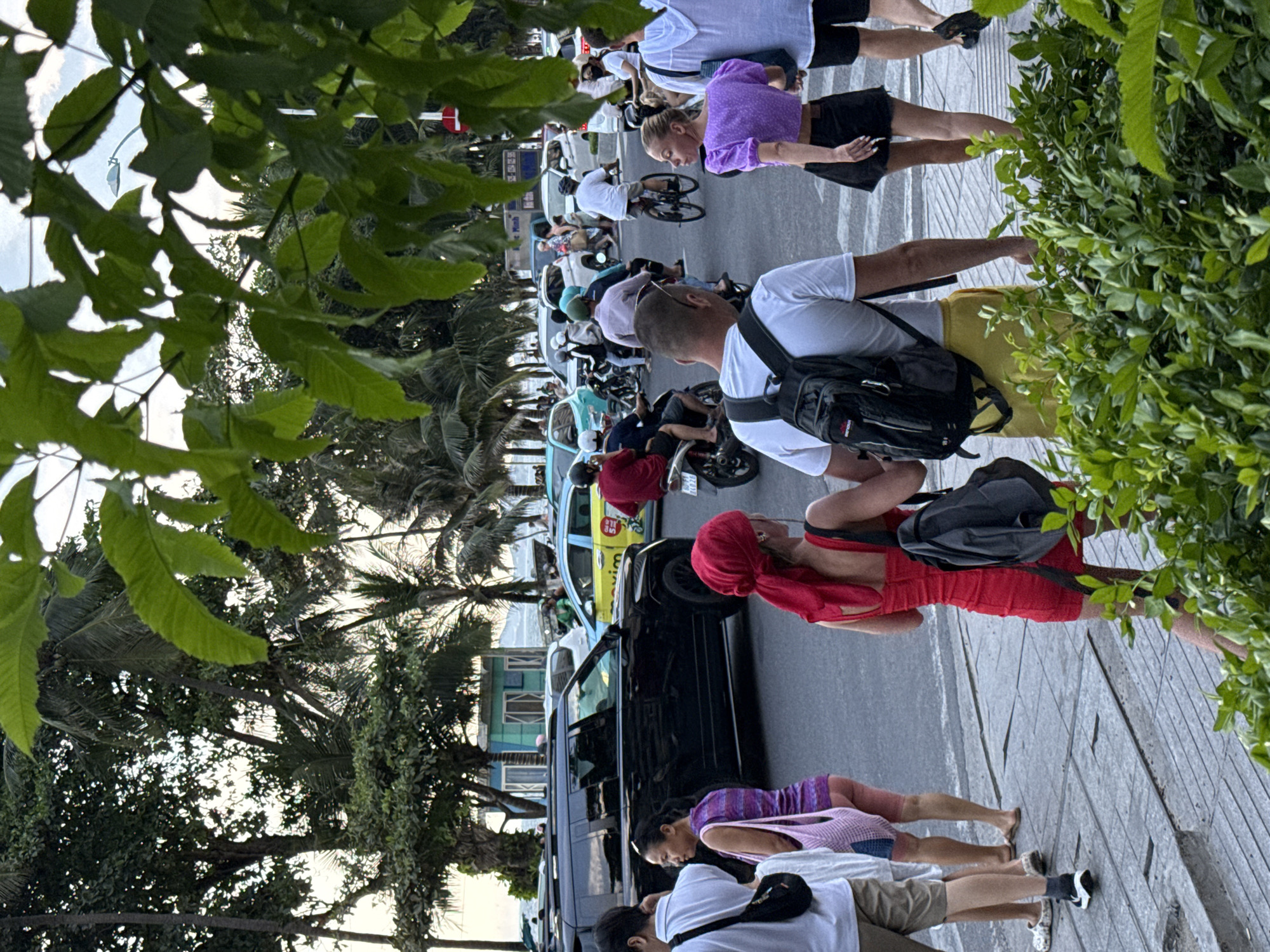 A street scene in Nha Trang Ward, Vietnam, depicts a busy intersection with various modes of transportation. A large black SUV is parked on the right side of the road, facing left. To its right, a yellow taxi with "Maxim" branding is visible, indicating a ride-sharing service. Several motorcycles are present, some carrying passengers. People are walking on a sidewalk in the foreground, with some crossing the street. The environment is characterized by lush green foliage framing the top and right edges of the image, suggesting a tropical or subtropical setting. The sky is overcast, and palm trees are visible in the background, further confirming the location's climate. The overall impression is one of a lively urban environment with a mix of pedestrian and vehicular traffic.