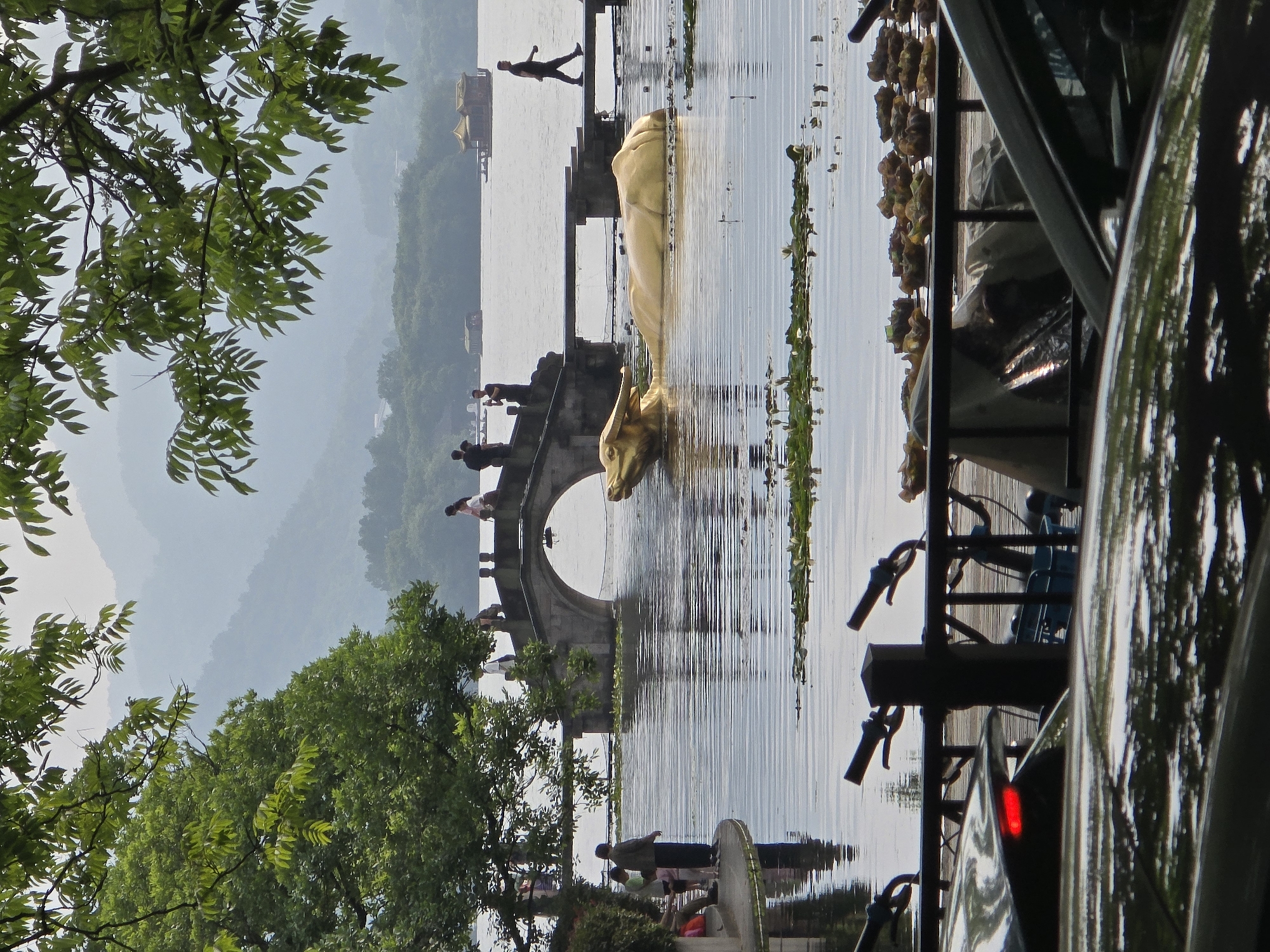 The image displays a picturesque waterside scene, likely a lake or large pond, set against a backdrop of misty mountains. The time of day appears to be daytime, with diffused light suggesting either an overcast sky or a hazy atmosphere.
In the foreground on the right, a bicycle rack holds multiple bicycles, with their handlebars and frames clearly visible. A dark vehicle, possibly a car, is partially visible, with a red taillight prominent. The top-left corner is framed by lush green tree branches and foliage, indicating a tree overhanging the view.
The midground features a calm body of water traversed by a stone arch bridge, on which several people are seen walking. A striking golden sculpture of a mythical animal, possibly a dragon or a Qilin, rises from the water near the bridge, its head and a large part of its body visible. Patches of aquatic vegetation float on the water's surface. More people are scattered along the distant shorelines and pathways, suggesting a public park or scenic area.
In the background, layers of green mountains or hills recede into a gentle haze, lending a sense of depth and tranquility to the landscape. A few small, traditional-looking structures are faintly discernible in the distance along the water's edge. The scene evokes a peaceful and culturally rich environment. No visible text is present in the image.