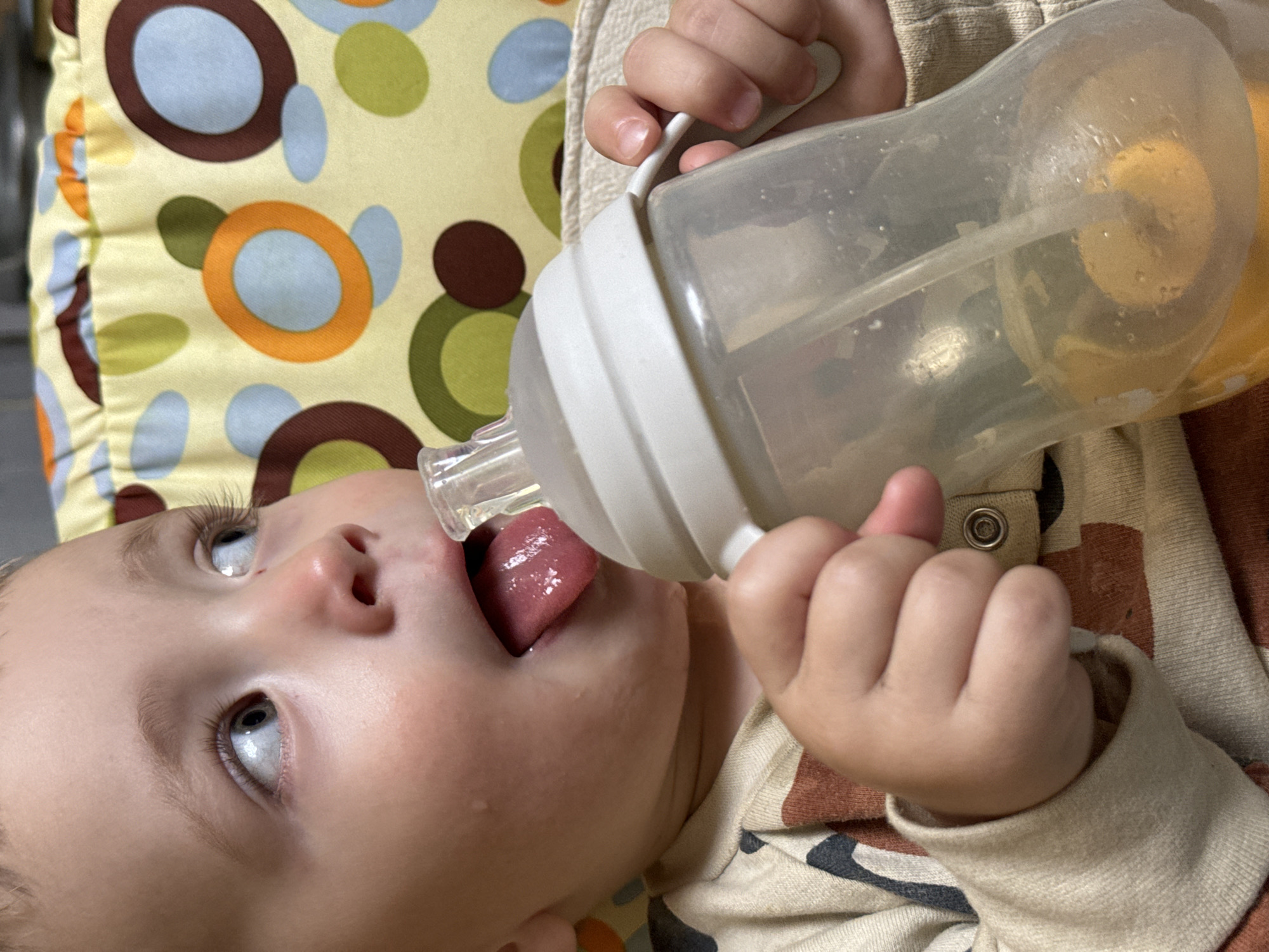 A close-up shot depicts a baby lying on a patterned surface, holding and drinking from a translucent bottle. The bottle, which appears to contain a yellow liquid, is held in the baby's hands, with the nipple positioned in the baby's mouth. The baby's tongue is visible, and there are droplets near their mouth. The surface underneath the baby features a pattern of circles in various colors, including blue, brown, green, and orange, against a yellow background. The location is Unknown, Unknown.