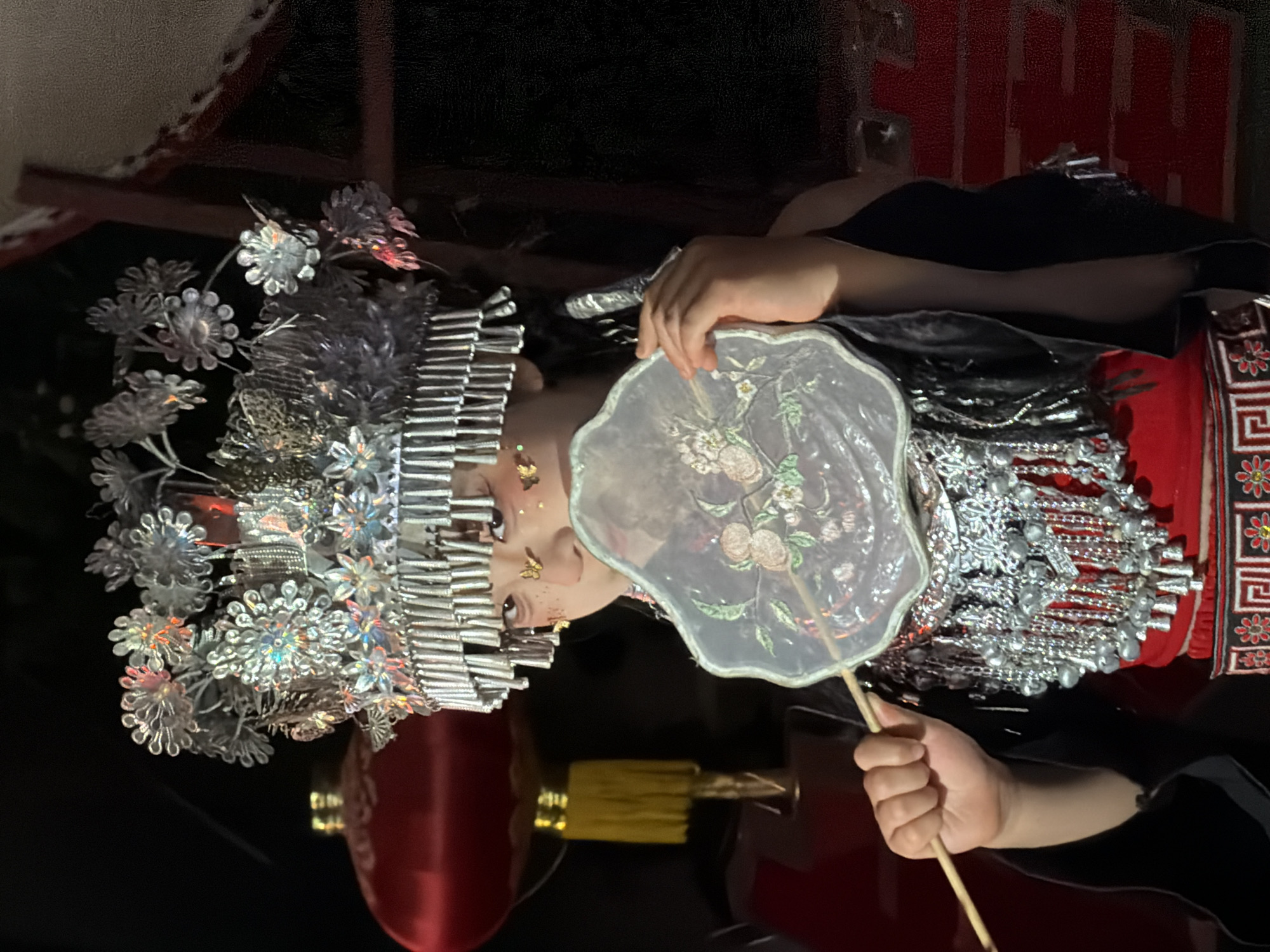 A young girl in traditional ethnic attire, likely from a minority group in China, is pictured in a dimly lit indoor setting. She wears an elaborate silver headdress adorned with floral motifs and hanging ornaments. Her face is partially obscured by a translucent fan, decorated with embroidered flowers. The fan is held by a thin wooden stick. She has makeup on her face, including what appear to be gold flecks or butterflies near her eyes and on her cheeks. Her outfit consists of a red garment, embellished with silver jewelry featuring numerous dangling bell-like ornaments and beaded chains. A black outer layer with reflective elements is visible. In the background, a large red lantern with a yellow tassel is partially visible, along with what seems to be a brick wall and dark fabric. The overall scene suggests a cultural performance or festival, possibly in Yuzhong District, China, given the context of traditional attire and symbolic decor.
