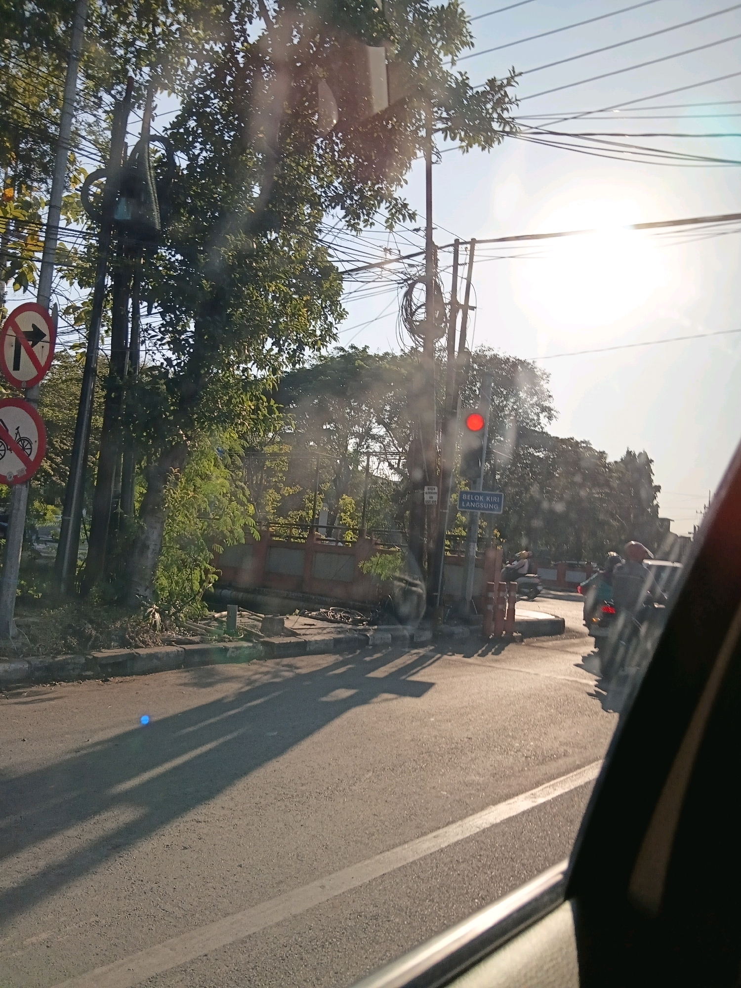The image depicts a street scene in Surabaya, Indonesia, captured from inside a vehicle. The perspective is looking outwards towards the right side of the street. A prominent traffic light with a red signal is visible, indicating a stop. Several traffic signs are present, including one with a red circle and a black arrow pointing right, signifying "no right turn." Another sign below it displays a bicycle within a red circle, indicating "no cycling." A blue street sign with white lettering reads "BELOK KIRI LANGSUNG," which translates to "Turn left directly."
The environment includes trees lining the street, overhead electrical wires, and buildings in the background. A wall with an orange-brown hue and decorative elements runs along the sidewalk. On the street, several motorcycles and a few individuals are visible, suggesting moderate traffic. The road surface is asphalt, and shadows of trees and possibly other objects are cast across it due to the sunlight. A small, bright blue light is reflected on the road. The overall lighting suggests it is daytime, with the sun casting long shadows. The presence of these specific street signs and the overall urban environment are characteristic of a street in Surabaya.