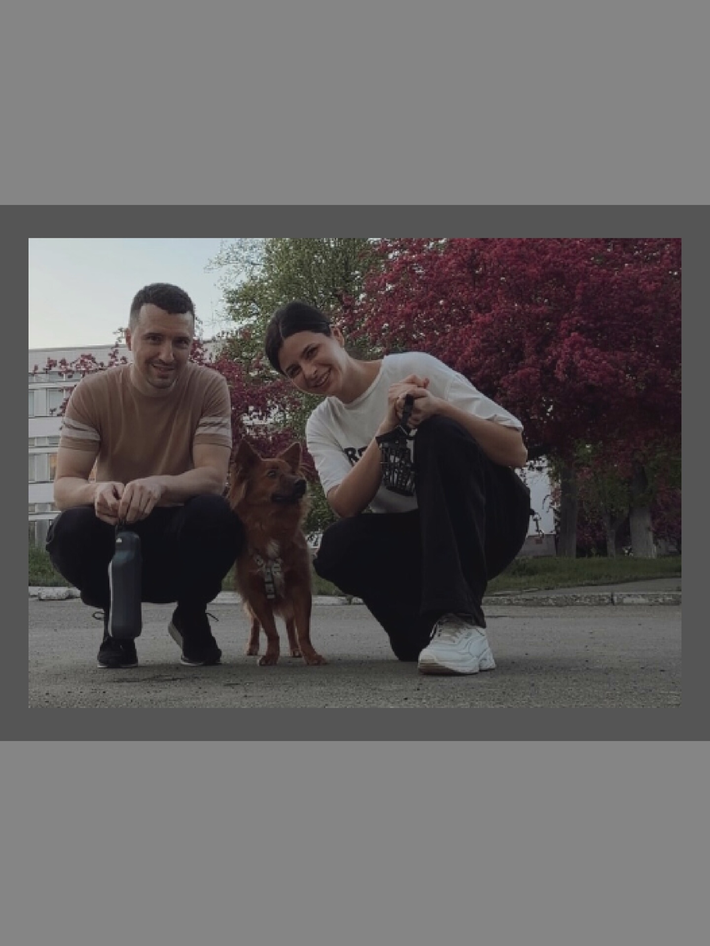 Two individuals, a man and a woman, are crouching on asphalt, with a small, reddish-brown dog standing between them. The man, on the left, wears a light brown t-shirt and dark pants, with a dark water bottle beside him. The woman, on the right, wears a white t-shirt and black pants, and is holding a camera. Behind them, a building with windows is visible, and a tree with vibrant red foliage. The scene appears to be outdoors, possibly in a park or residential area.