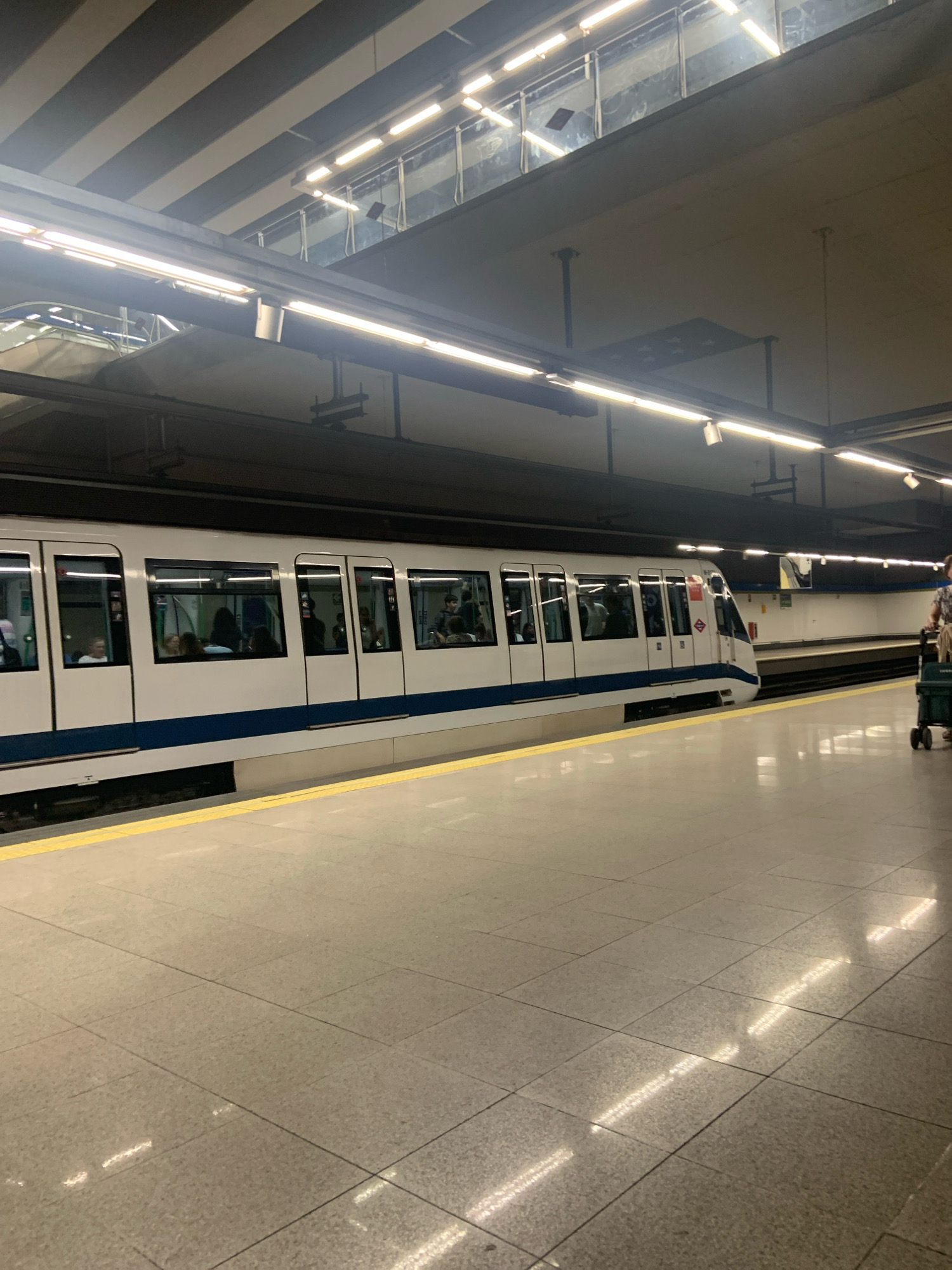An interior shot depicts a white and blue metro train stopped at a platform in Alcorcón, Spain. The train has large windows, and several passengers are visible inside. The platform is made of gray tiles and has a yellow safety line along the edge. Overhead, fluorescent lights illuminate the scene, reflecting on the polished floor. In the background, a person is seen with a shopping cart. The ceiling is constructed with beams and panels, typical of a subway station.
