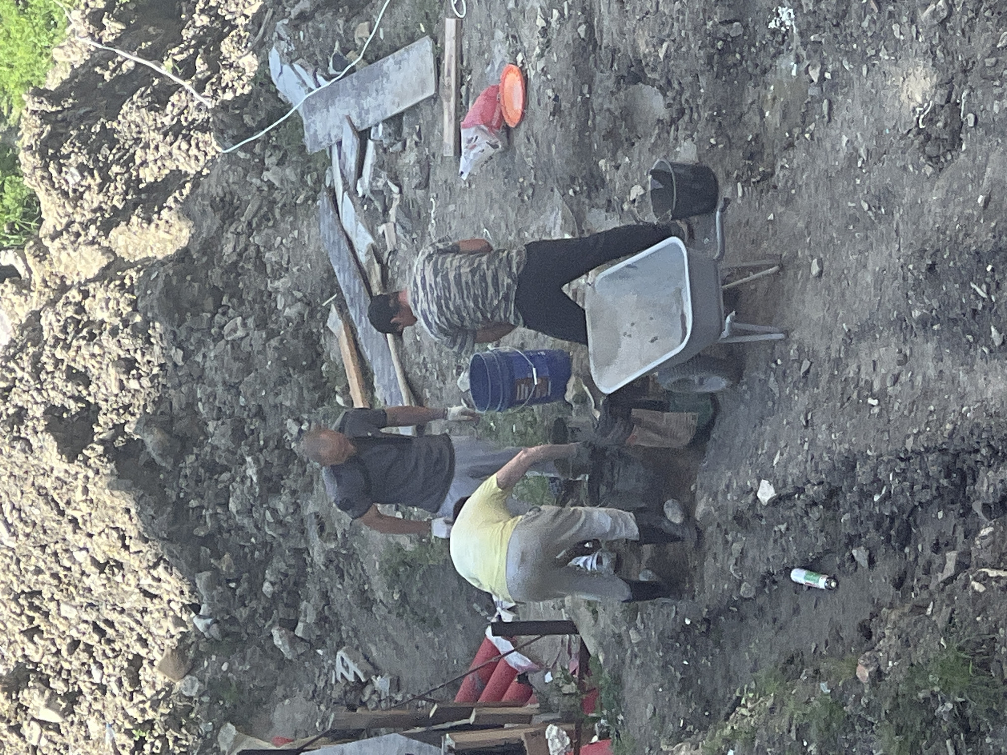 This image, captured in Sochi, Russia, depicts an outdoor scene featuring three adult males engaged in manual labor on uneven, muddy ground interspersed with construction debris.
**Environment:** The ground is predominantly dirt and mud, with numerous scattered rocks, gravel, and fragments of concrete. In the background, a substantial mound of excavated earth and rock is visible. Patches of green vegetation are present in the upper right and lower left portions of the frame. Various wooden planks and boards are spread across the mid-ground and background, alongside discarded materials. A thin white cable or rope runs diagonally from the upper right toward the center. The scene is illuminated by natural daylight.
**Personnel and Actions:**
1. **Individual 1 (Left-Center):** A male, seen from behind, is wearing a light yellow t-shirt, light-colored pants, and dark rubber boots. He is bent over, actively working with a dark container on the ground, possibly mixing materials with a tool whose handle is visible.
2. **Individual 2 (Center-Right):** A male, positioned behind Individual 1, is wearing a dark grey t-shirt, light grey pants, and white work gloves. He stands upright, looking down at the work area, and holds a blue plastic bucket in his left hand, poised above the container where Individual 1 is working.
3. **Individual 3 (Right):** A male, also seen from behind, is wearing a camouflage-patterned t-shirt and dark pants. He stands upright to the right of the work area, facing partially left, observing the activity.
**Objects:**
* **Equipment:** A grey metal wheelbarrow is positioned in the lower mid-ground, to the right of Individual 1. Two dark plastic buckets are on the ground: one near Individual 1's work area and another to the right of the wheelbarrow. The blue plastic bucket is held by Individual 2.
* **Debris/Materials:** Red cylindrical objects, possibly pipes, are stacked against a structure on the far left. A white plastic bag containing red material, alongside an orange plastic plate, is lying on the ground in the mid-ground right. A white and green aerosol can or similar container is on the ground in the lower left foreground. Assorted wooden planks, some appearing aged, are scattered throughout the background.