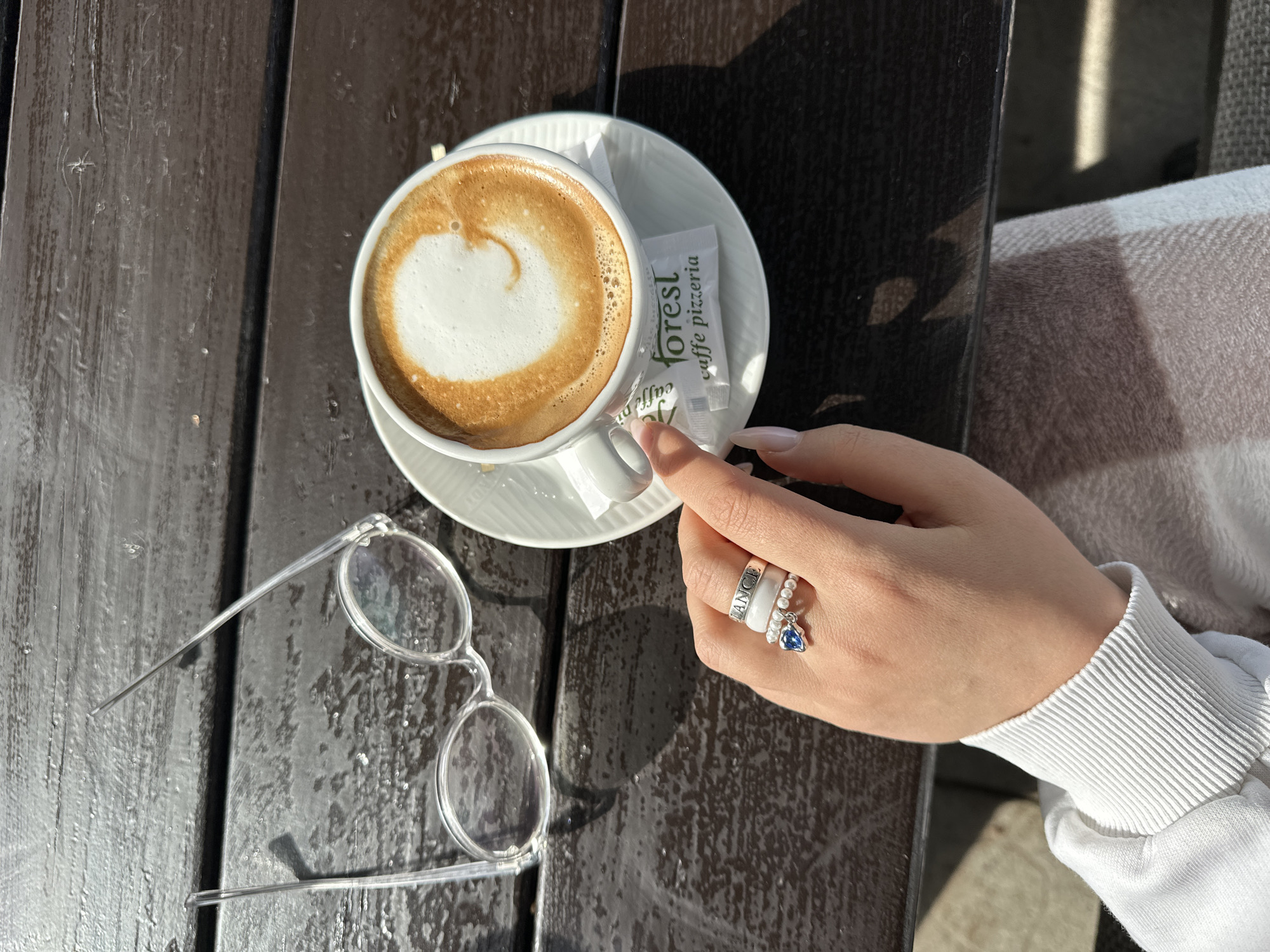 A close-up shot captures a moment at an outdoor café, likely in Bar, Montenegro, evidenced by the "forest caffe pizzeria" sugar packet. A cup of cappuccino with a frothy heart design sits on a white saucer. A woman's hand is poised near the cup, seemingly about to add sugar. The woman wears rings, including one with a blue stone. Clear-rimmed glasses are on the wooden table alongside the coffee cup. The table is outdoors, likely on a patio or terrace. A white sweater sleeve is visible in the frame's right edge.