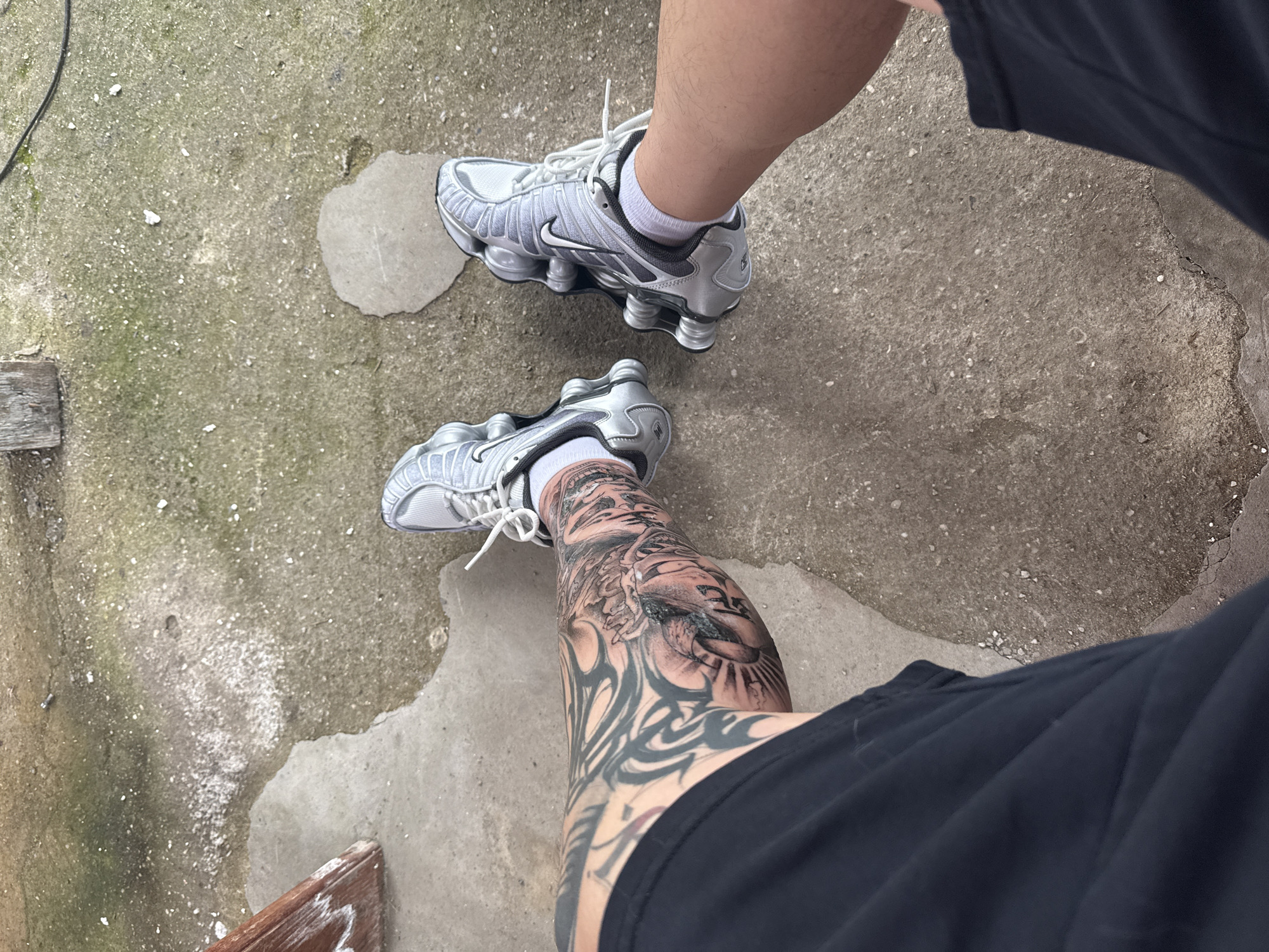 The image shows a person standing on a concrete surface. They are wearing a pair of silver Nike Shox TL sneakers with white socks and black shorts. The person's left leg is heavily tattooed. The concrete surface appears to be outdoors, possibly a patio or sidewalk. It is difficult to discern the exact location in Burgas, Bulgaria, without additional information or landmarks in the image.