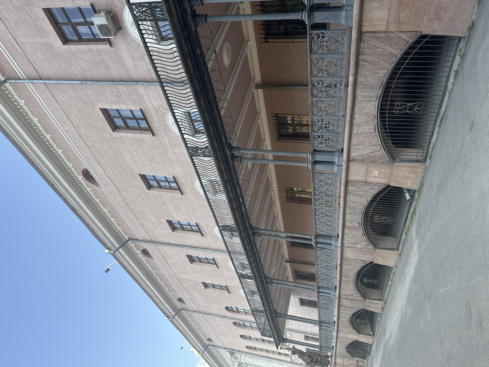 The image shows the exterior of a light pink, multi-story building with a long, covered balcony running along the second floor. The balcony features an ornate, black metal railing with a wave-like pattern and is supported by dark grey pillars. Below the balcony, the building's foundation is constructed with rough-hewn pinkish-brown stone, featuring arched openings covered by black metal grates. The ground in front of the building is a paved, grey asphalt surface. The sky above is clear and blue. Architectural details like decorative cornices and multiple windows with dark frames are visible on the building's facade. A statue of a person in dark clothing is partially visible on the far left of the image, near the base of the building. The building appears to be located in a city environment, given the paved road and urban architecture. The overall scene suggests a historical or grand architectural style.