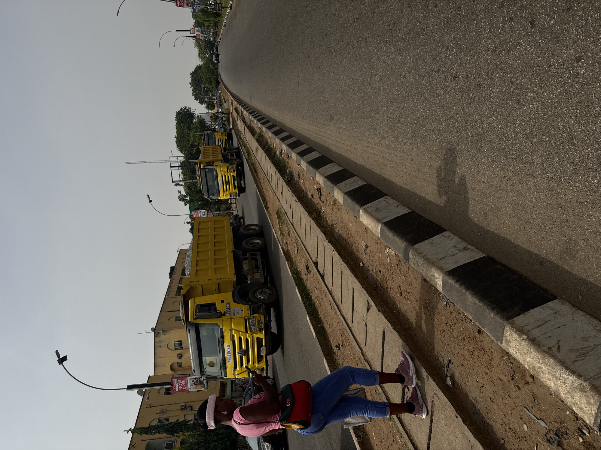 The image displays a daytime street scene in Abuja, Nigeria, featuring a paved road extending into the distance on the right. A concrete median, painted with alternating black and white stripes, separates the road from a dirt shoulder and a concrete drainage ditch.
A person, wearing blue jeans, a pink short-sleeved top, pink sneakers, and a pink headband, is standing on a paved area next to the dirt shoulder in the foreground. They carry a red, green, and yellow shoulder bag and appear to be holding a phone in their right hand. The person's shadow is cast distinctly on the asphalt, indicating bright, possibly hazy, daylight conditions.
A line of large yellow dump trucks is parked along the side of the road to the left of the person. The closest truck prominently features the name "ZEBERCED" on its cab and "CONSTRUCTION GROUP" below it, with the number "2027" visible on its dump bed. A dark-colored car is partially visible behind the person and the first truck.
In the background, light-colored multi-story buildings and numerous green trees are visible along the street. Streetlights with distinctive curved poles extend over the roadside. Various signs and billboards are scattered in the background; one near the trucks clearly displays "SPAR NOW OPEN" and "KASCO". The sky above is bright but appears somewhat hazy. The scene depicts a typical urban or semi-urban environment with ongoing activity or parked construction vehicles.