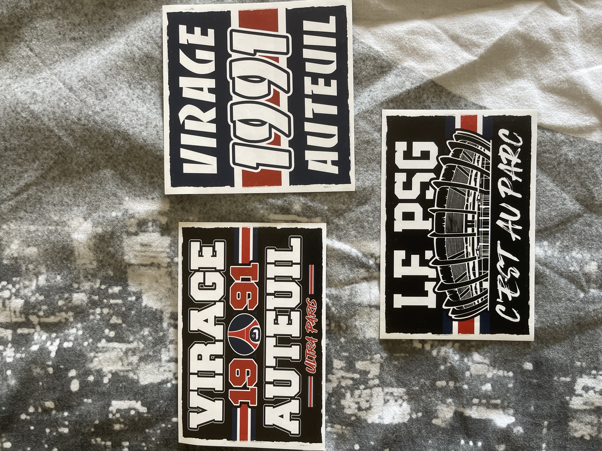 The image displays three stickers with the text "VIRAGE 1991 AUTEUIL" and "LE PSG C'EST AU PARC". The stickers are laid out on a gray, textured surface. The text on the stickers refers to the Parc des Princes stadium in Paris, France, which is the home stadium of the Paris Saint-Germain Football Club. The stadium is depicted on one of the stickers.
