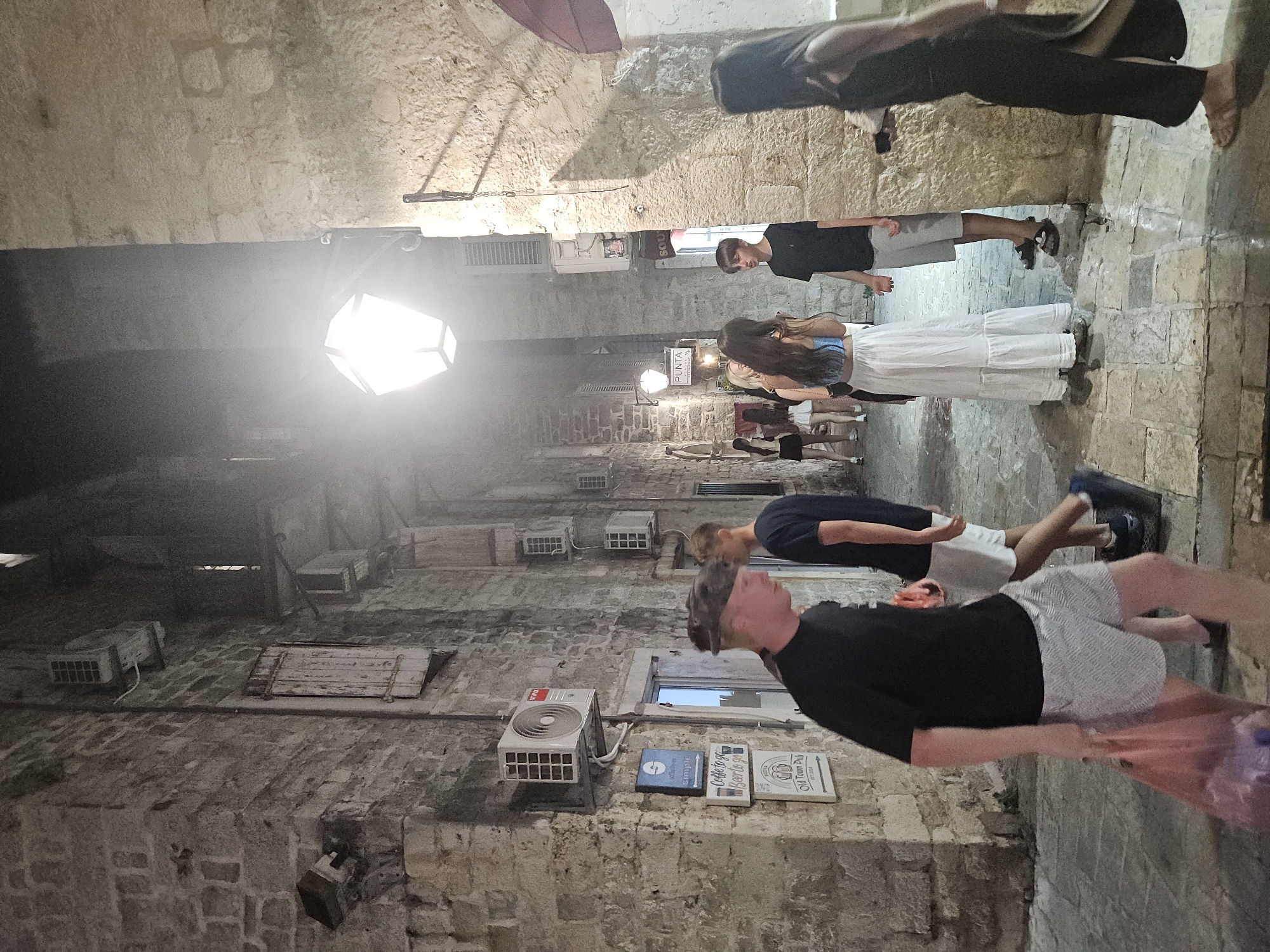 In Budva, Montenegro, a narrow cobblestone alleyway is illuminated by a large, faceted lantern overhead and a smaller one further down. The stone walls of the buildings are ancient, with signs indicating "PUNTA" and "Old Town Pub." Several air conditioning units are mounted on the walls. A group of young people walk through the alleyway at night. In the foreground, a man wearing a black t-shirt and patterned shorts carries a pink plastic bag filled with items. Behind him, another man in a black t-shirt and grey shorts walks away. Further into the alley, a woman in a white maxi skirt and blue top stands with a group of friends. A woman in a black outfit walks towards the viewer on the right side of the image. The lighting creates long shadows and a somewhat hazy atmosphere.