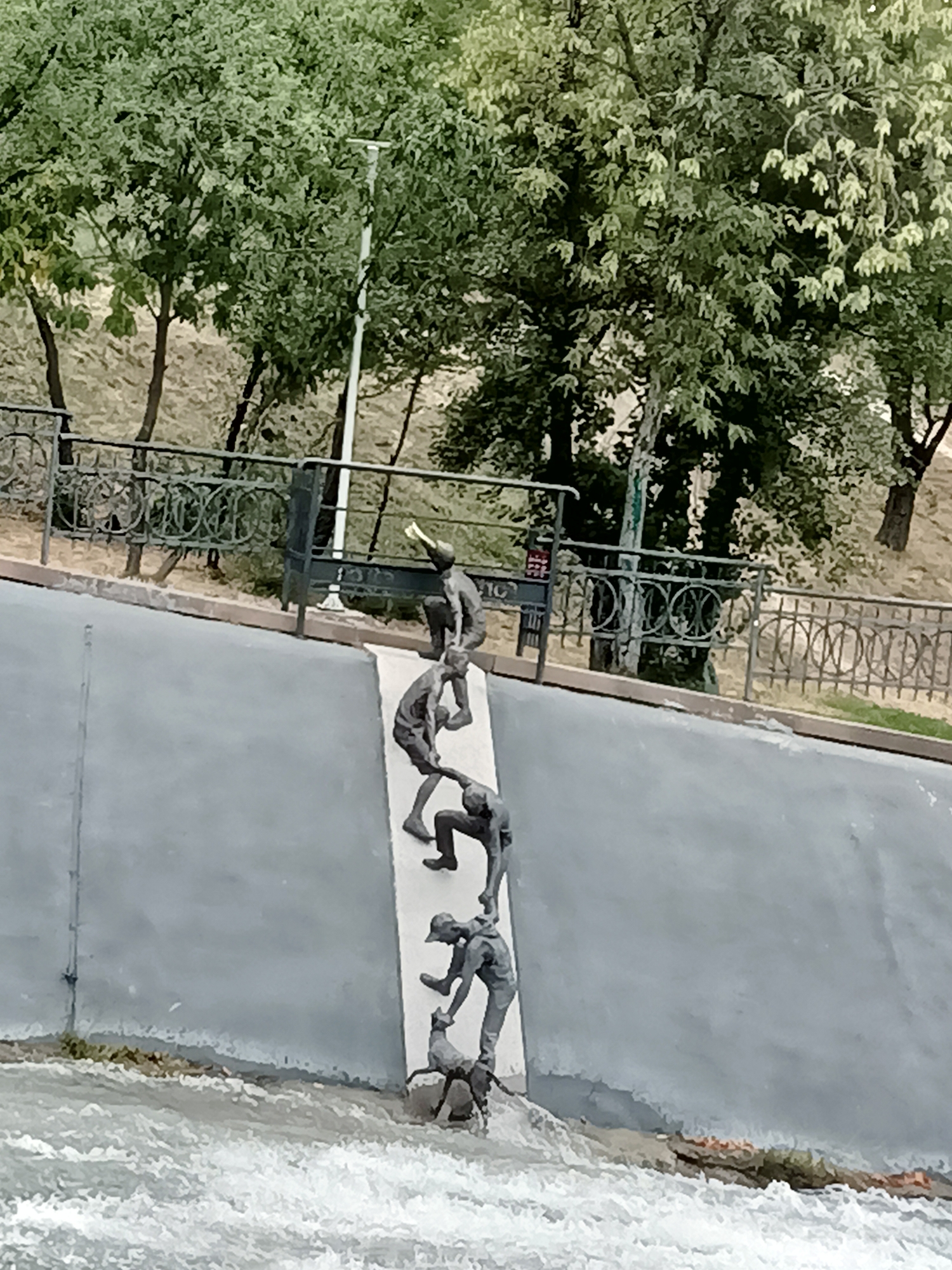 The image captures a sculpture depicting a group of people helping each other up a steep incline, possibly representing teamwork or overcoming adversity. The sculpture is located in the city of Almaty, Kazakhstan, along the embankment of the Malaya Almatinka River. The scene is set against a backdrop of lush green trees and a metal fence. The water in the river is turbulent, suggesting a strong current or rapids. The overall impression is one of dynamic action and human connection, with the sculptures conveying a sense of struggle and mutual support.