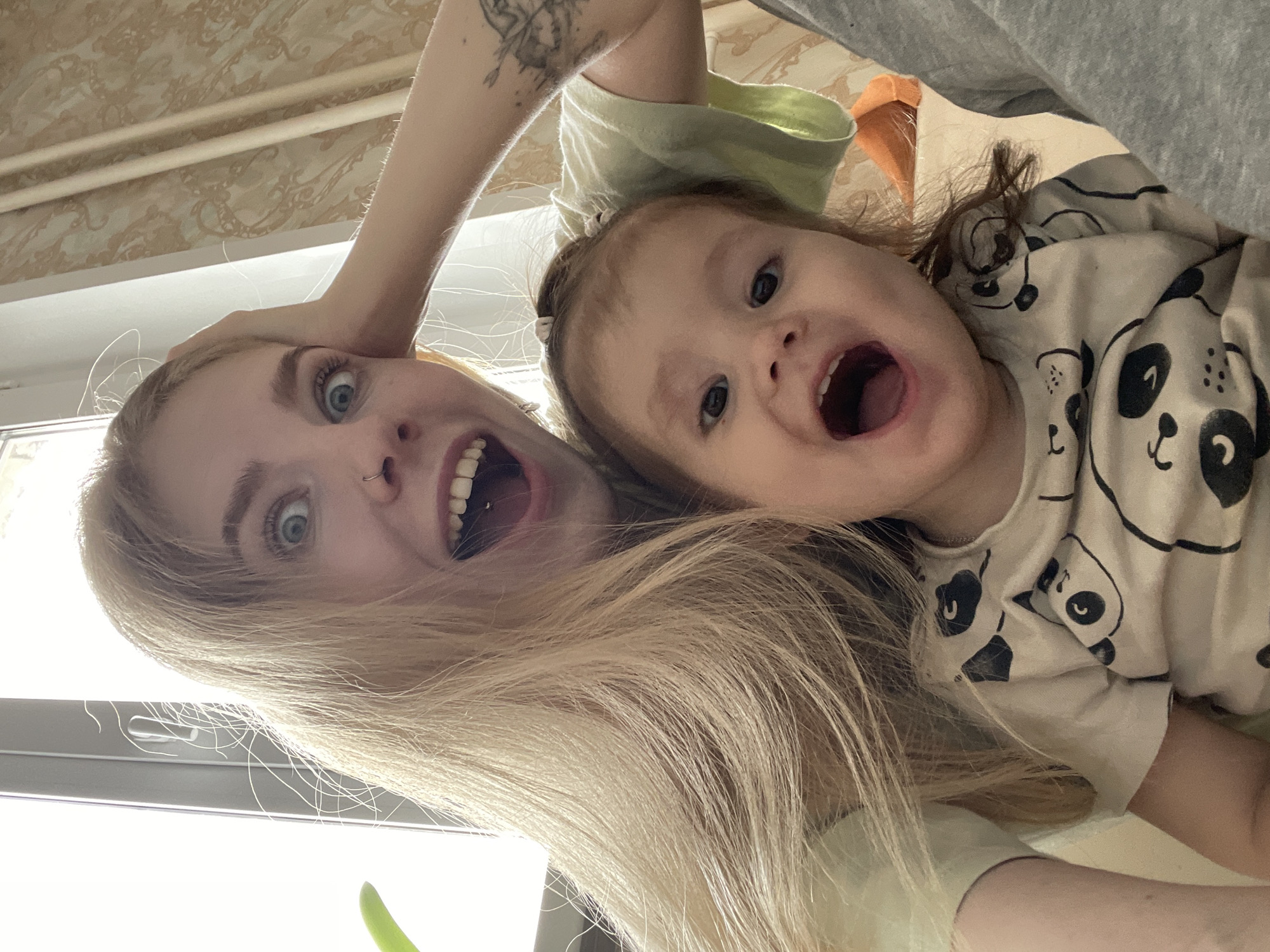 The image displays a playful, low-angle perspective of an adult female and a young child, positioned overhead and looking directly into the camera, creating a selfie-like shot. Both individuals have wide-open mouths and expressive eyes, conveying feelings of joy, excitement, or playful surprise.
The adult female has long, flowing blonde hair, bright blue eyes, and a nose piercing. A tattoo, depicting an intricate design of what appears to be a winged figure or mythical creature, is visible on her right upper arm. She is wearing a light-colored, possibly light green, top.
Beside her, a young child, likely a toddler, with light brown hair partially tied back, is wearing a cream-colored top featuring multiple black and white cartoon panda faces.
The scene is indoors, with a patterned ceiling or wallpaper visible in the background, adorned with ornate, swirling motifs. A bright light source, likely a window, casts light onto the subjects from the left. The overall atmosphere is cheerful and dynamic. No discernible text is visible within the image. The location context for this media file is Izhevsk, Russia.