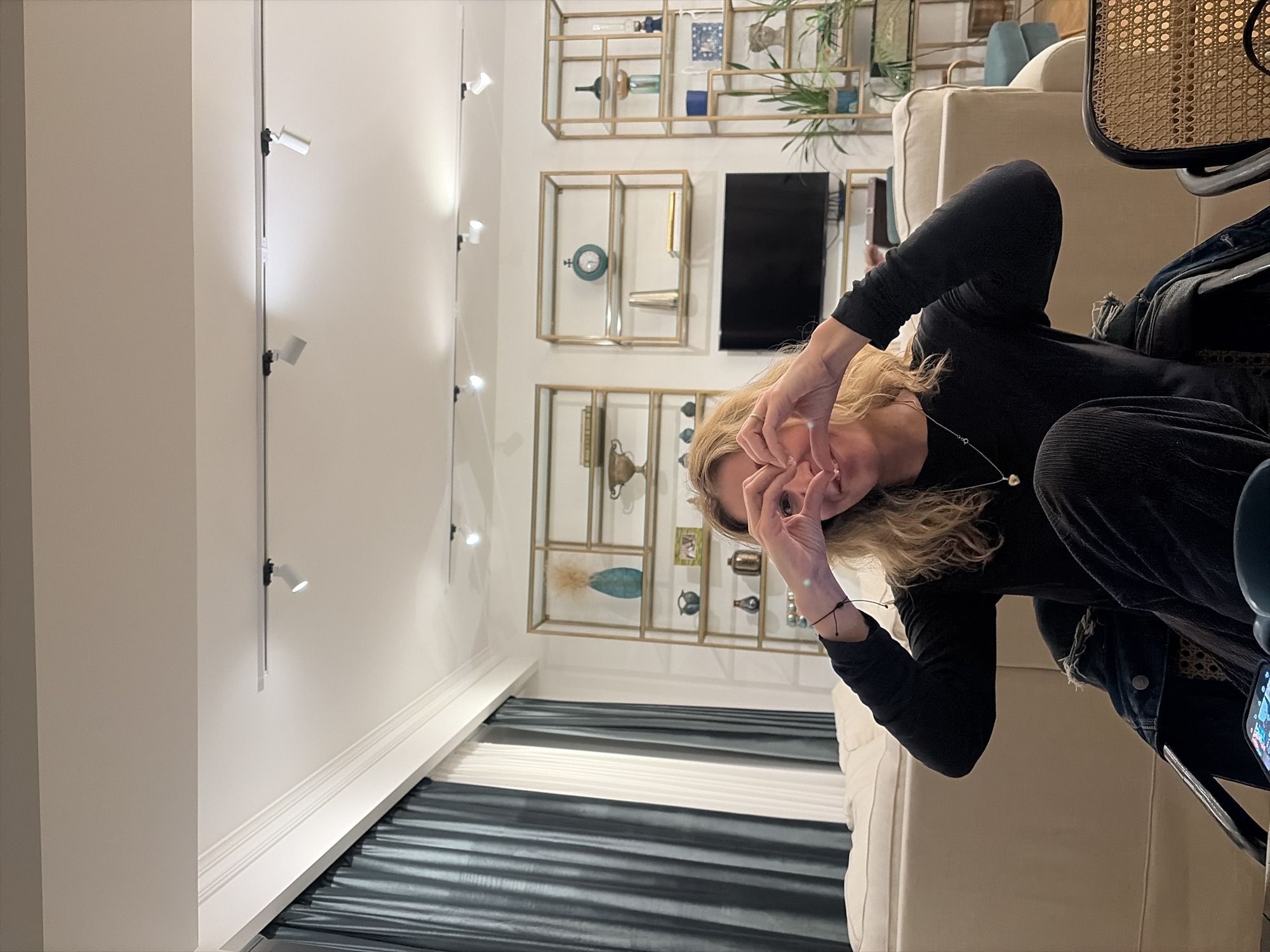 A woman with light blonde hair is seated on a light-colored couch. She is wearing a black long-sleeved shirt and dark trousers, and has a bracelet on her left wrist. She is making a circle shape with her fingers in front of her face. In the background, there are shelves with decorative items and a television. The walls are white, and there is a track lighting system on the ceiling. The window is covered with blue and white striped curtains. Based on the interior design, this image likely was taken in Tbilisi, Georgia.