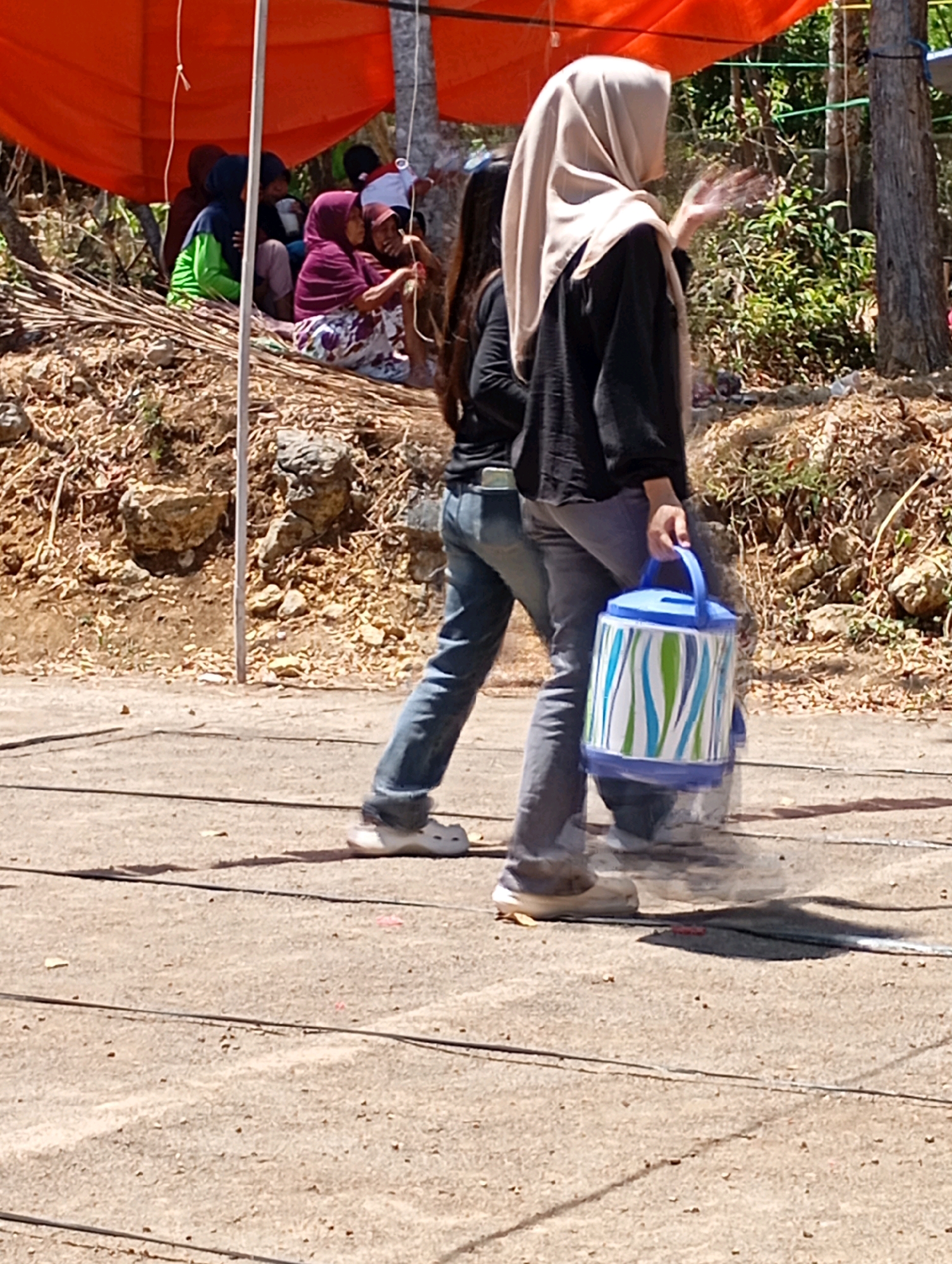 In Ketepung, Indonesia, a woman wearing a beige hijab and a black long-sleeved shirt walks on a dirt path, carrying a blue and white cylindrical container with green accents. She is accompanied by another person whose legs are visible, wearing blue jeans. In the background, several individuals are seated under an orange canopy, some of whom are wearing headscarves. The environment is outdoors, with trees and dry vegetation visible, suggesting a rural or semi-rural setting.