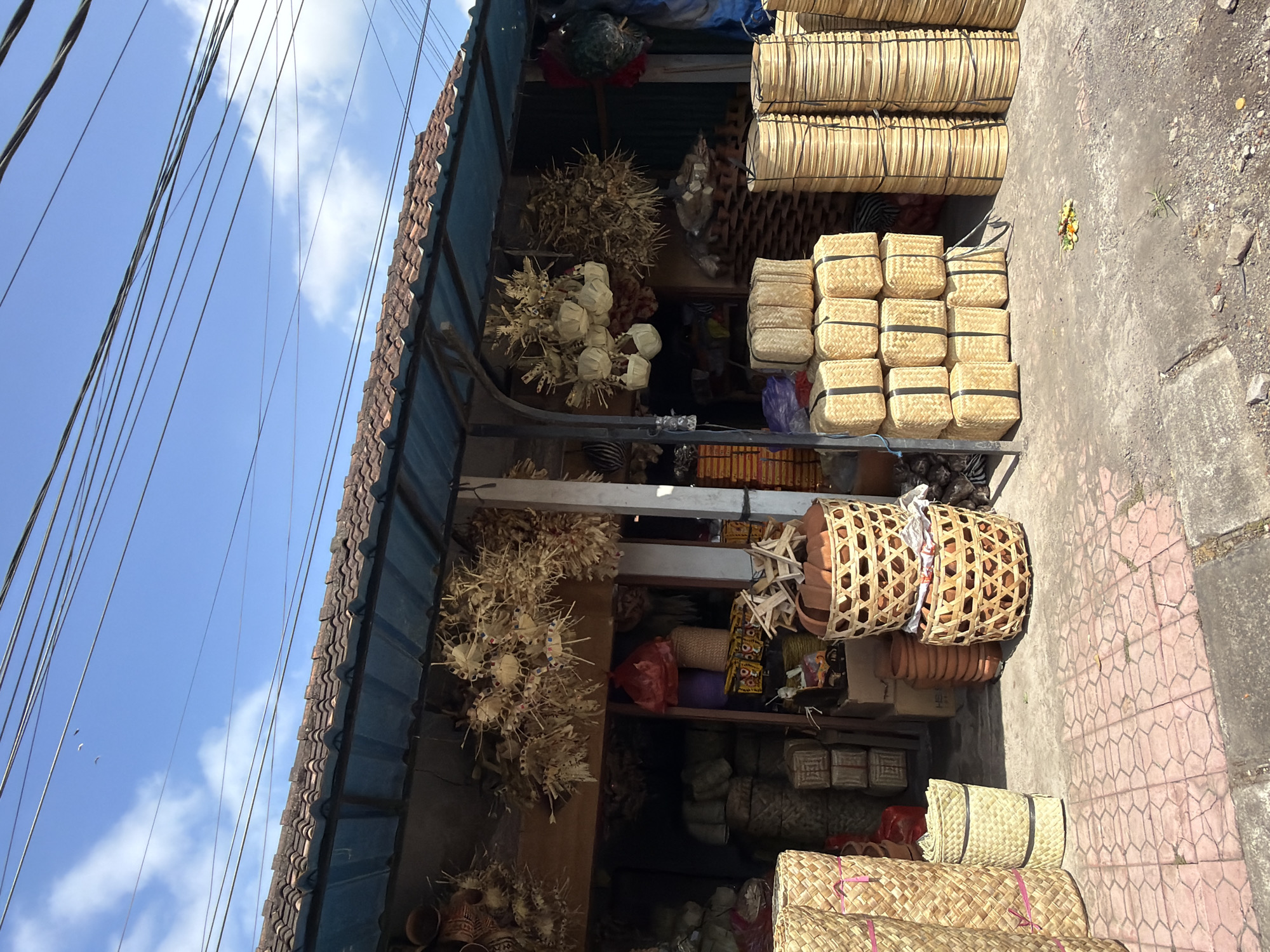 This is a frontal, eye-level, medium shot of a roadside market stall selling woven bamboo products in Blahbatuh, Indonesia. The stall is open to the street, with a tiled roof supported by metal poles. Hanging from the overhang are numerous dried woven items, possibly decorative or functional. In front of the stall, the ground is paved with reddish-pink hexagonal tiles, and beyond that, the ground is a mix of concrete and dirt with small stones and debris.
Various sizes and shapes of woven bamboo baskets and containers are displayed. To the left, there are tall, cylindrical baskets. In the center, stacked rectangular baskets with black straps are prominent, along with a couple of larger, circular woven baskets. To the right, stacks of thin, circular woven items, possibly plates or lids, are piled high. Behind the merchandise, the interior of the stall is visible, showing more stacked baskets and some bags, including a red one.
The sky above is bright blue with scattered white clouds. Several thin black electrical wires crisscross the upper portion of the frame. There are no people visible in the image, nor are there any apparent actions or interactions taking place. The scene appears to be a typical street-side vendor in a warm, sunny climate.