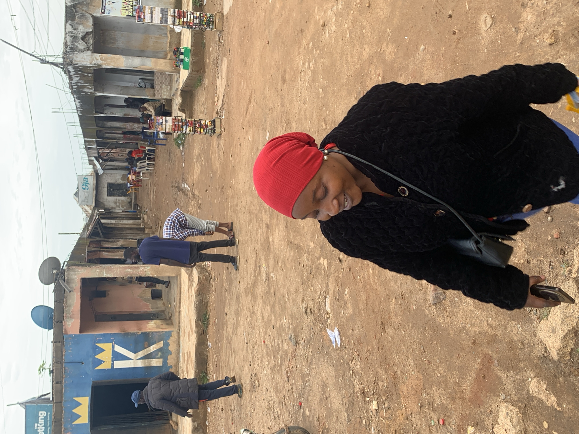 This image captures an outdoor street scene in an urban or semi-urban area within Jos, Nigeria. In the immediate foreground, a woman of African descent is seen in motion, her head turned slightly to the left, exhibiting a smile with her eyes partially closed. She wears a red headscarf, a textured black long-sleesleeved jacket with gold-colored buttons, and blue trousers. A small black shoulder bag is visible on her right side, and she holds a dark-colored smartphone in her right hand. Her left ear features a pearl-like earring.
The ground surface consists of reddish-brown, unpaved dirt, interspersed with small stones and scattered debris, including white paper and small red circular objects. To the left of the woman, a man wearing a dark blue cap, a dark puffer jacket, and dark trousers is walking away from the viewer, towards a building. This building is painted blue and features prominent yellow signage, including a crown emblem and the letter "K," indicative of a "BetKing" establishment (partially visible "etKing" text). Two large satellite dishes are affixed to the roof of this building. Another person is partially visible within its open doorway.
In the midground-right, a man in a blue t-shirt and dark trousers stands facing slightly to his left. Near him, another man, dressed in a light-colored plaid shirt and light trousers, is bent over, facing away from the viewer. Two vertical market displays, each constructed of multiple stacked tiers of small, colorful packaged goods, are prominently positioned on the right side of the street. Adjacent to these displays, a green crate containing bottles or cans rests on a raised concrete platform.
Further into the background, the unpaved thoroughfare continues, bordered by additional single-story structures. These buildings show signs of aging, including peeling paint, exposed rebar on rooflines, and a visible "9ja" sign on one rooftop. Several individuals are seated at plastic tables (red, yellow, blue) and chairs, suggesting an outdoor communal or dining area. Overhead, utility wires are visible against an overcast sky, indicating diffuse natural light. The scene depicts an active street with modest commercial and residential infrastructure.