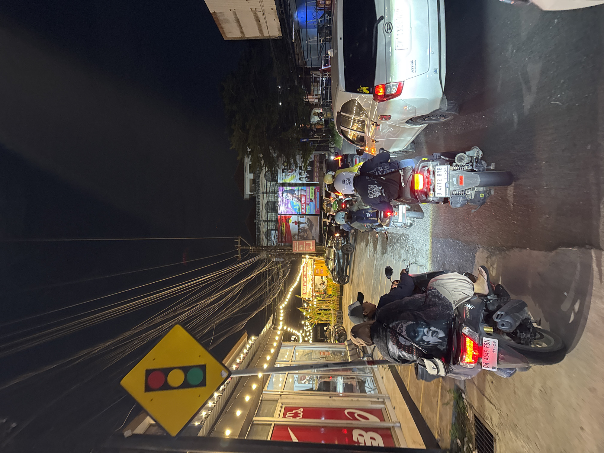 The image depicts a traffic scene at night on a city street in Denpasar, Indonesia. A traffic light sign is visible on the left side of the frame, indicating a yellow background with red, yellow, and green circles representing the traffic signals. The street is lined with businesses featuring illuminated storefronts and string lights. Several motorbikes are present, with riders wearing helmets and casual clothing. Some of the motorbikes have passengers. One motorbike in the foreground displays a license plate reading "DK 4546 FBN" with a date "11-29". A white car is also visible in traffic, with a license plate that is partially obscured. The sky is dark, suggesting nighttime. The overall impression is one of a busy urban environment with moderate traffic congestion.