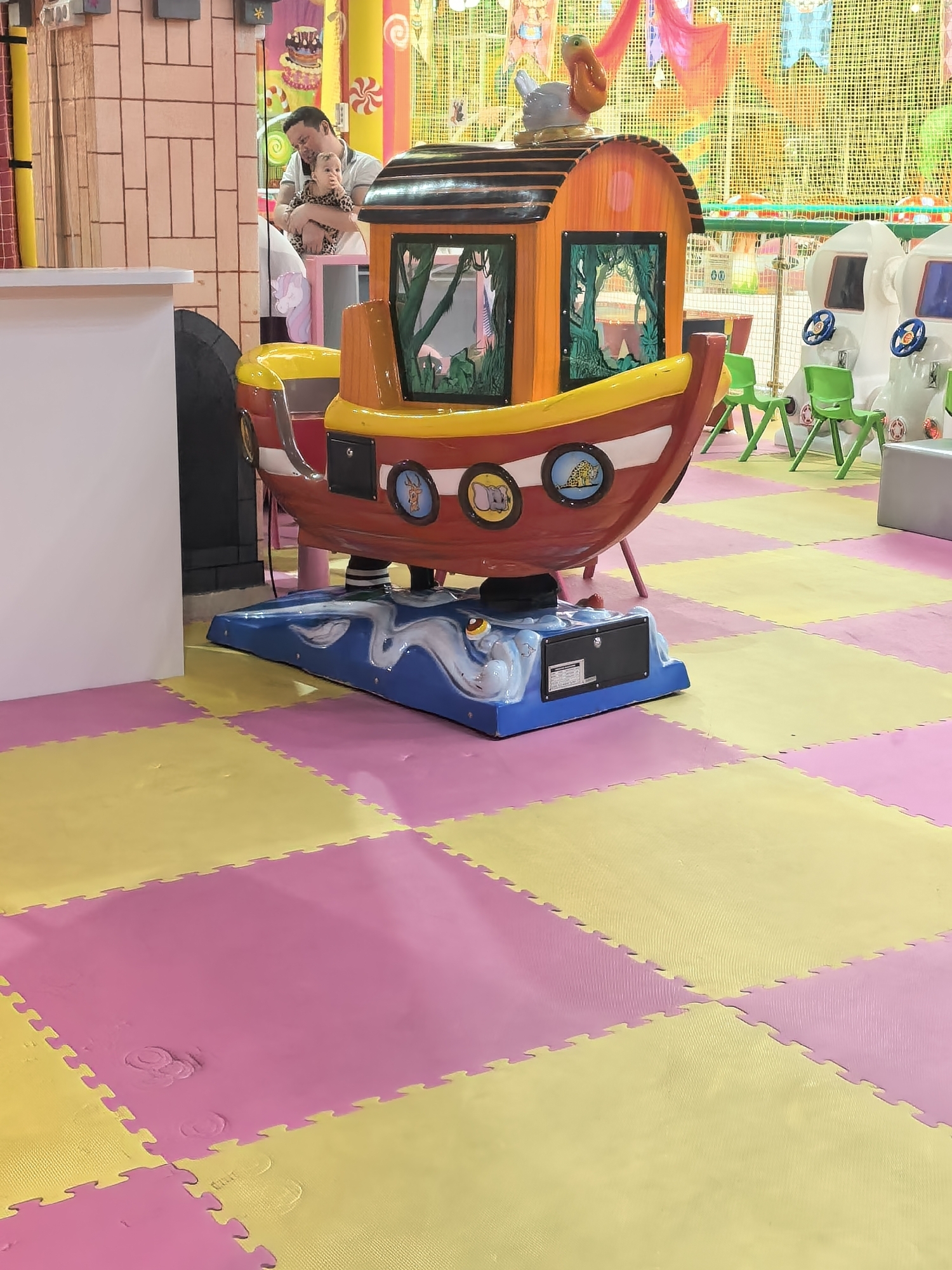 The image shows a colorful children's play area, possibly indoors. The flooring is made of interlocking foam mats in alternating pink and yellow squares. A ride shaped like a boat is in the center, featuring a red and yellow hull, an orange cabin, and a blue base decorated with cartoonish waves. A man is holding a baby in the background near a play structure designed to look like a building with a turret. Other children's rides (rocket ships) and green chairs are visible in the background. The location is Sterlitamak, Russia.