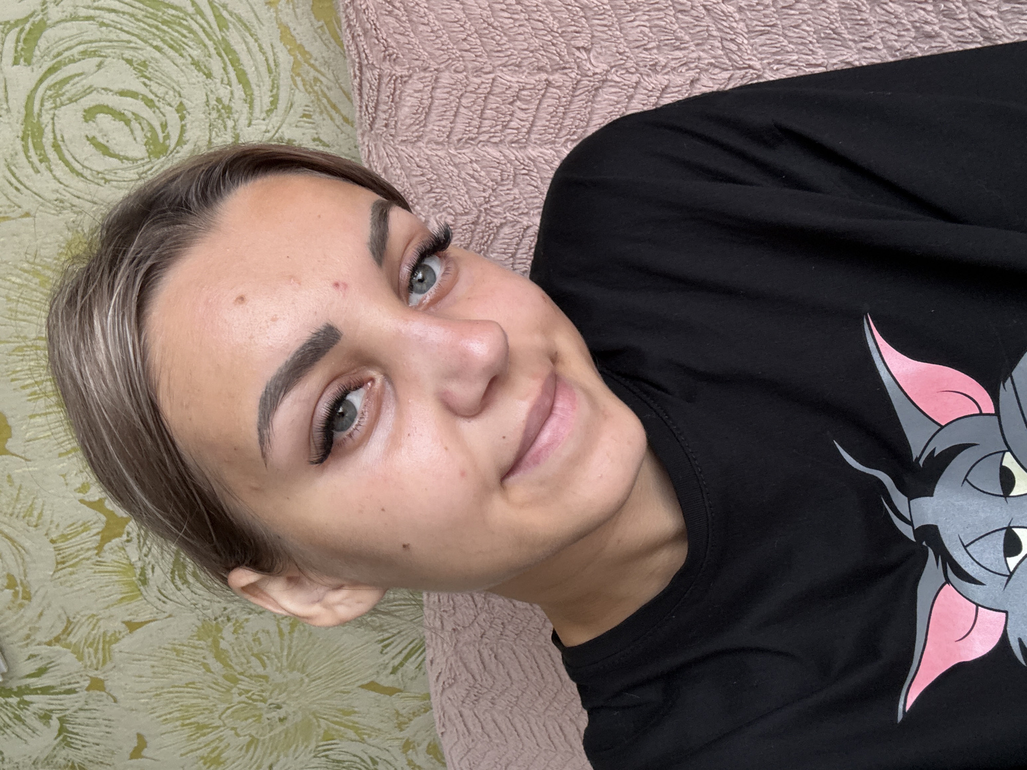 A close-up selfie taken from a slightly elevated angle shows a young woman with fair skin and light brown hair pulled back from her face. She is wearing a black t-shirt with a cartoon character graphic of Tom from Tom and Jerry. Her eyes are blue-green, and she has prominent eyelashes and eyebrows. There are a few blemishes visible on her forehead. She is smiling faintly, and her head is tilted slightly to her left. The background consists of a textured wallpaper with a floral or abstract pattern in green and yellow tones, and a pink textured surface, possibly a sofa or blanket, is visible behind her. The lighting appears to be natural, coming from the front. There is no indication of the location being Krasnogorskoye, Russia.
