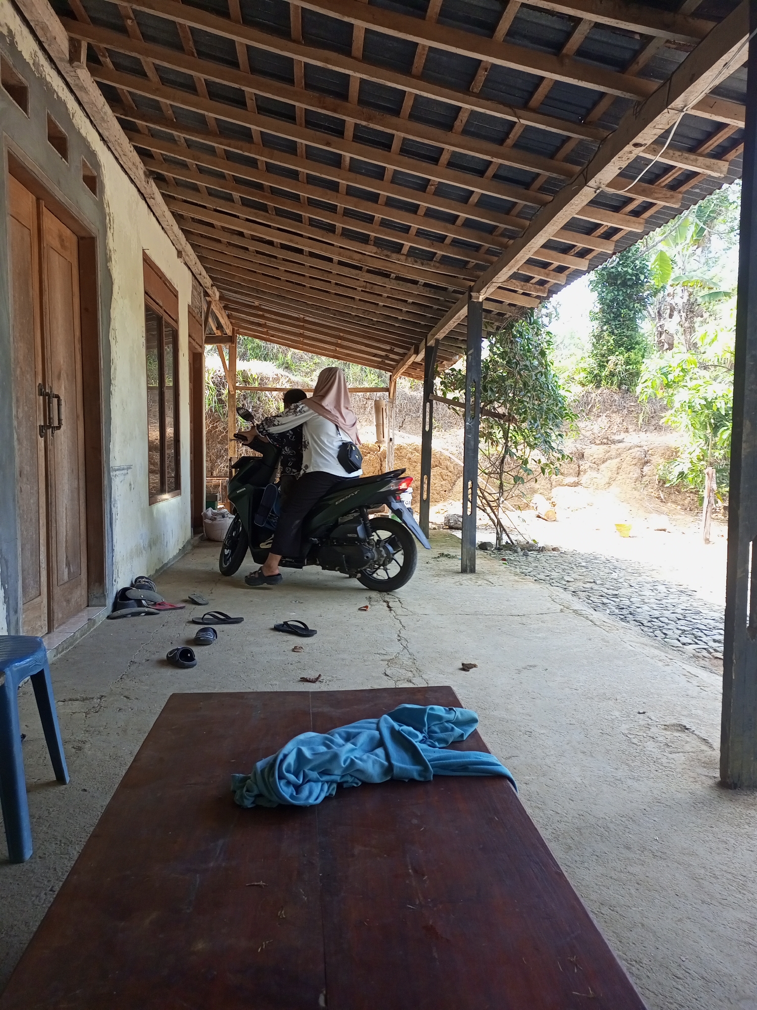 In Jatipurno, Indonesia, two individuals are on a dark green motorcycle parked on a concrete porch. The person in the back wears a light brown hijab and a white and black patterned shirt, while the person in front wears a white shirt with similar patterns and dark pants. Scattered on the porch are several pairs of sandals and flip-flops. To the left of the motorcycle, a blue plastic chair is visible. In the foreground, a dark wooden table has a crumpled light blue cloth on it. The porch is covered by a wooden-framed roof with exposed wooden beams and dark roofing material. Beyond the porch, the environment consists of a gently sloping, dry, dusty terrain with scattered vegetation and trees. A cobblestone path is visible extending from the porch into the yard. The scene appears to be a residential setting during daylight hours, characterized by its rustic construction and outdoor elements.
