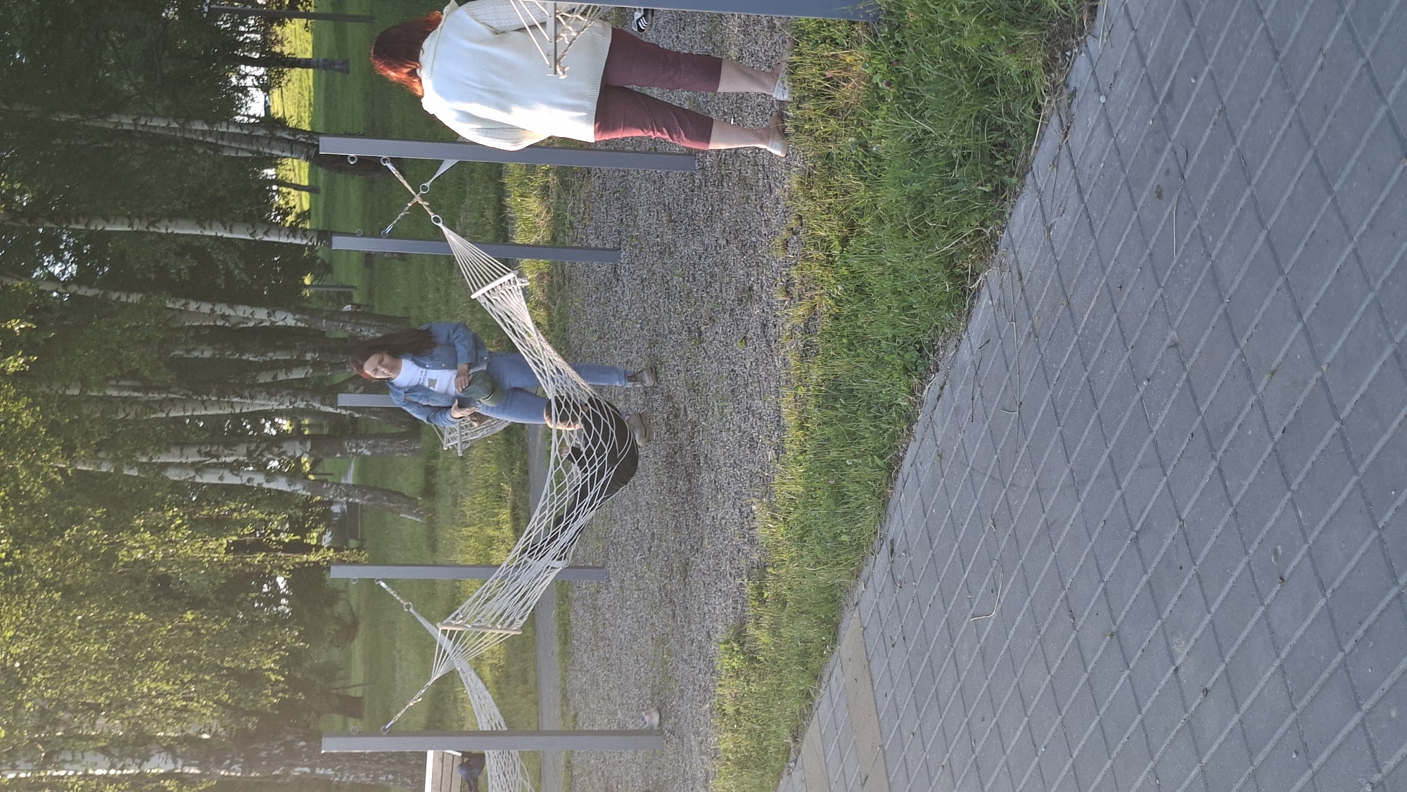 In the city of Smolevichi, Belarus, on a sunny day, two women are observed in a park area featuring hammocks. The woman on the left, wearing a denim jacket and jeans, stands beside a hammock, holding a dark green cap. A person is lying in the hammock. The woman on the right, seen from the back, wears a light-colored jacket and dark red cropped pants. She stands near another hammock. The environment includes birch trees with green foliage, gravel ground, and a paved walkway in the foreground.