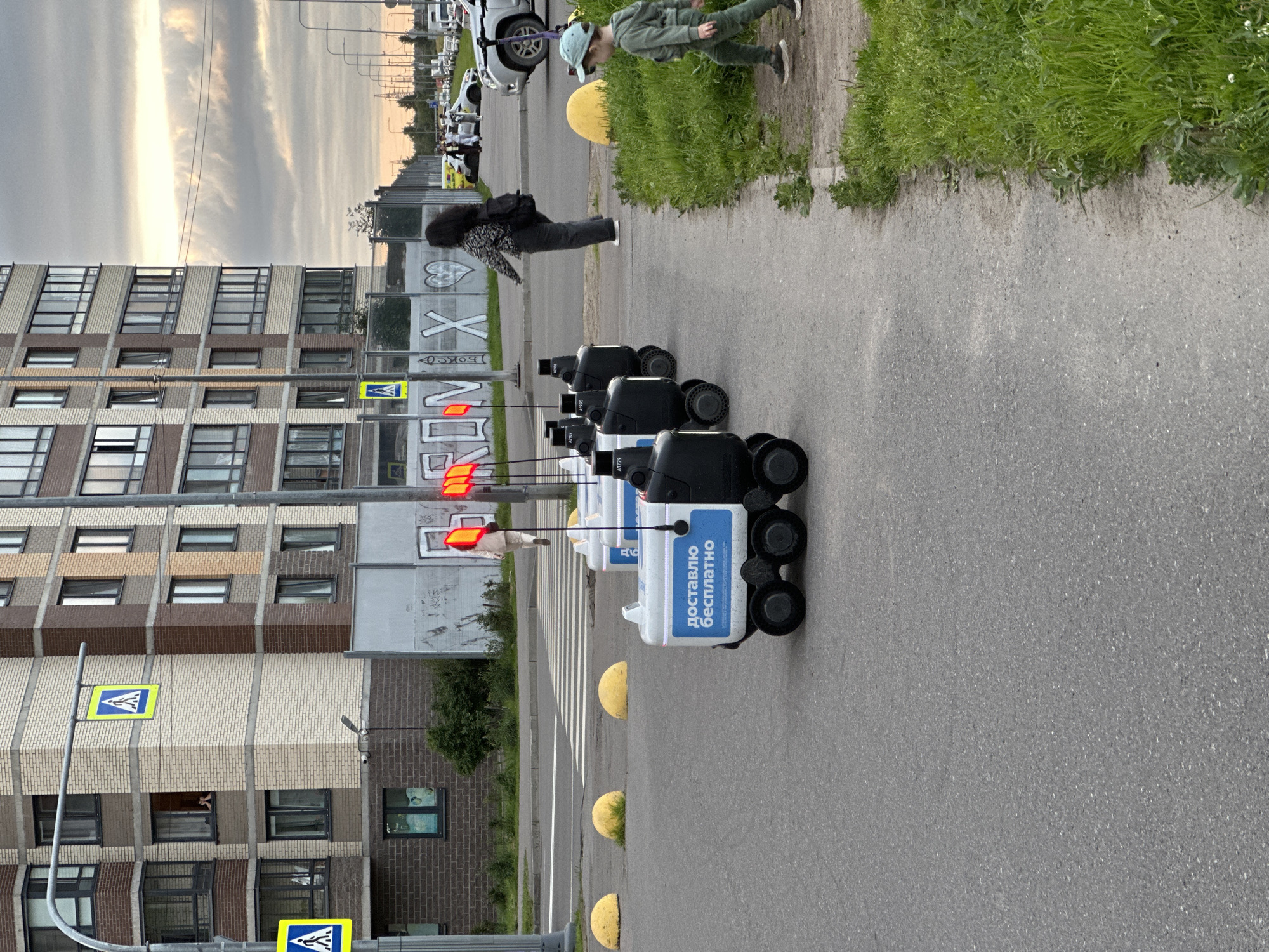 The image shows a street scene in Sinyavino, Russia, with a modern residential building in the background. In the foreground, a row of autonomous delivery robots are parked on the asphalt. The robots are white and black and have red indicator lights. One of the robots has Russian text that translates to "delivering for free". A child wearing a green hooded tracksuit and a light blue cap is walking on the grassy verge next to the sidewalk. A woman in a patterned top and dark pants with a backpack is walking away from the robots towards the street where several cars are parked. A pedestrian crossing is visible in the background, along with streetlights and some graffiti on a fence. The sky is overcast.