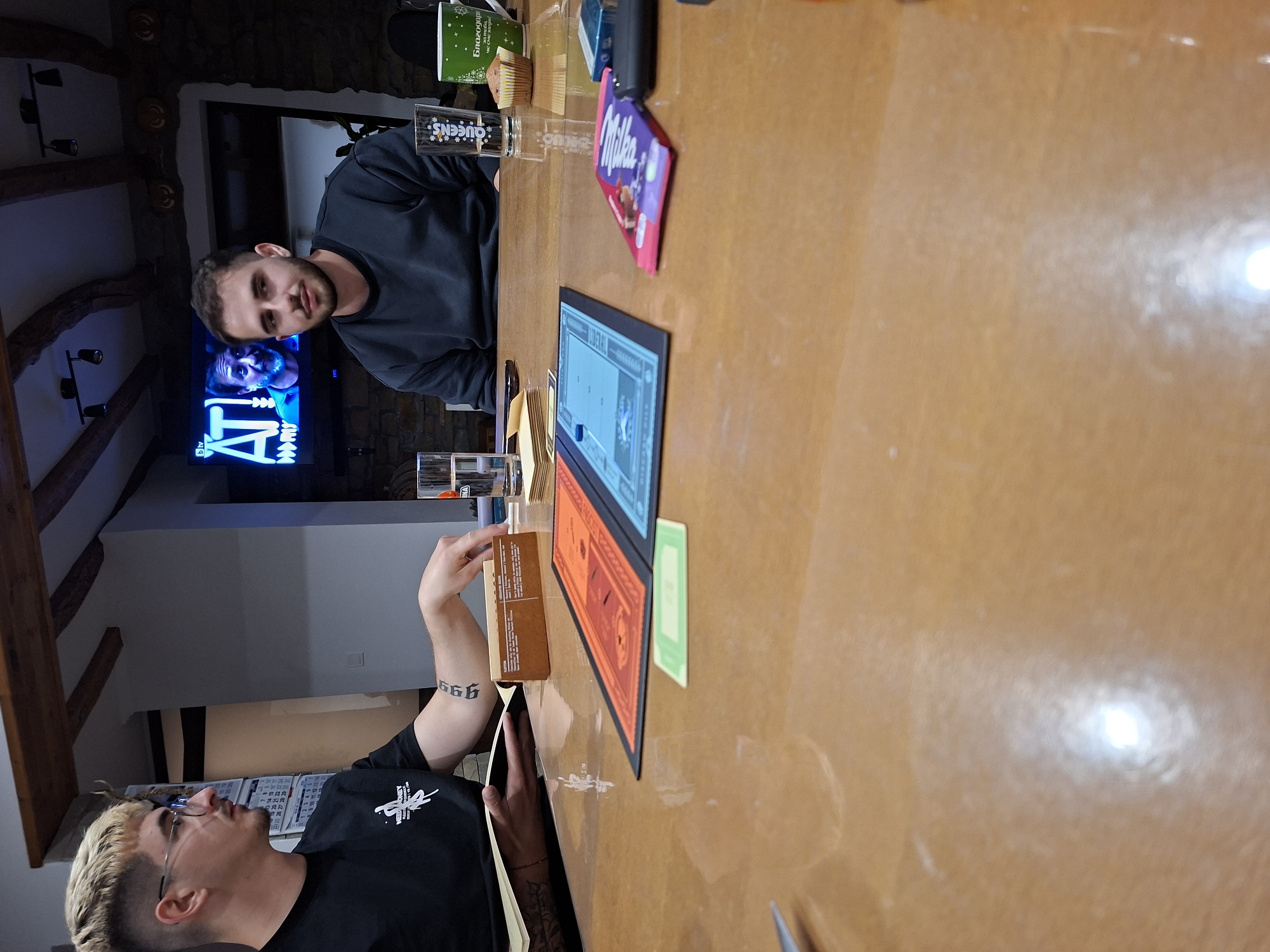 Two men are sitting at a table playing a board game. The man on the left is wearing glasses and a black t-shirt and has a tattoo on his arm. The man on the right is wearing a black sweatshirt. There is a board game, a box, cards, a Milka chocolate bar, a glass of matches, and a muffin on the table. In the background, there is a TV showing a program. The location is Gabrovo, Bulgaria.