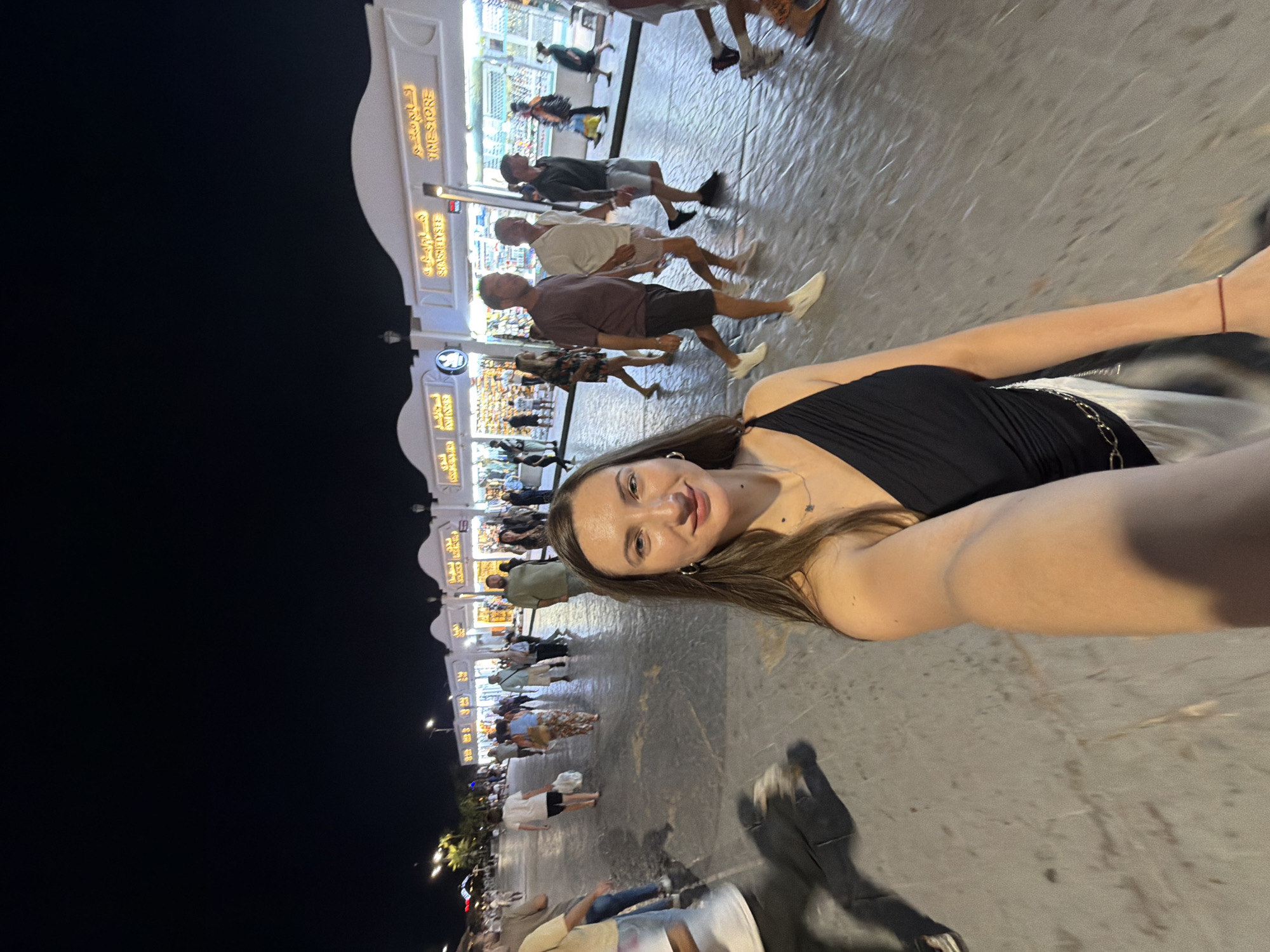 A woman is taking a selfie at night on a busy street in Sharm El Sheikh, Egypt. She is wearing a black one-shoulder top and a white skirt, with a gold chain belt. She has long brown hair and is wearing a necklace and earrings. Behind her, a street is lined with shops with illuminated signs, featuring Arabic and English text, indicating retail establishments such as "TIME STORE" and "SAKS FIFTH AVENUE". Several people are walking on the street, some are carrying shopping bags, and others are strolling. The street is paved and appears wet, reflecting the lights from the shops and streetlights. The sky is dark, suggesting it is nighttime. The overall scene depicts a vibrant commercial area in a tourist destination.