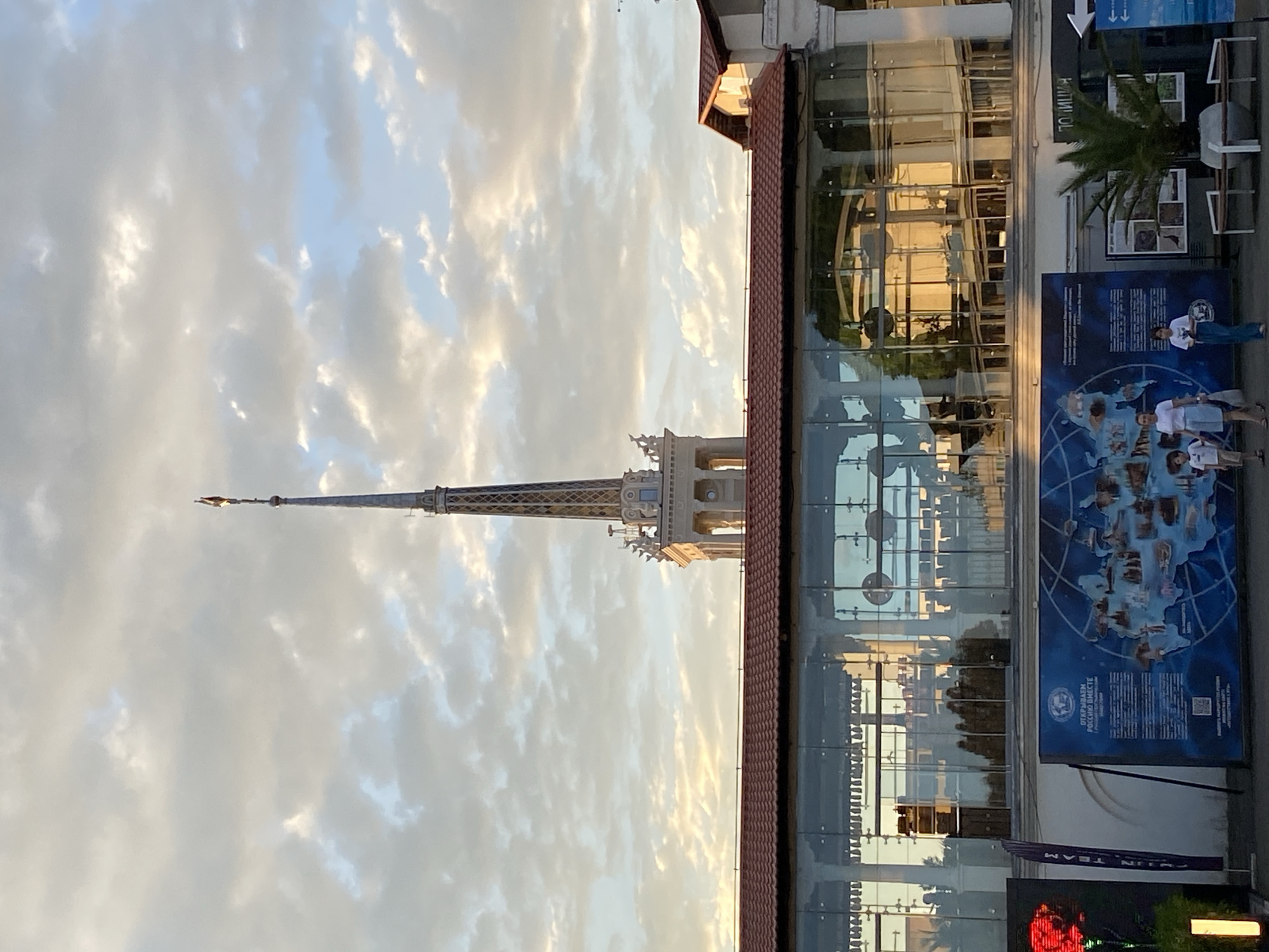 This is a photograph taken outdoors in Sochi, Russia, likely during the late afternoon or early evening, as indicated by the warm, diffused light and soft shadows. The sky is filled with scattered clouds, illuminated by the setting sun, creating a dramatic and somewhat ethereal atmosphere.
The main subject of the image is a tall, ornate spire that resembles a miniature Eiffel Tower. It is a slender structure with intricate latticework and decorative elements near its base, culminating in a sharp point. The spire stands against the cloudy sky.
To the right of the spire, a modern building with a large glass facade dominates the scene. The glass reflects the sky and the interior of the building, hinting at an indoor space with artificial lighting. A red-tiled roofline is visible along the top of this building.
In front of the glass building, there are several large posters or banners. One prominent blue banner displays a world map with various points of interest highlighted and text in Russian, including the phrase "ОТКРЫВАЕМ РОССИЮ ВМЕСТЕ" which translates to "WE ARE DISCOVERING RUSSIA TOGETHER". Another poster to the right features images of people and text, also in Russian.
A few people are visible near the posters, appearing to be tourists or visitors. They are casually dressed, suggesting a relaxed atmosphere. One group of three people, two women and a child, are standing near the blue banner, looking at it. Another person, a woman, is standing further to the right.
There is also a palm tree visible to the right of the building, adding a tropical touch to the scene, consistent with the location of Sochi. The overall impression is one of a public space, possibly an exhibition area or a waterfront promenade, with a blend of historical architectural style (the spire) and modern design (the glass building).