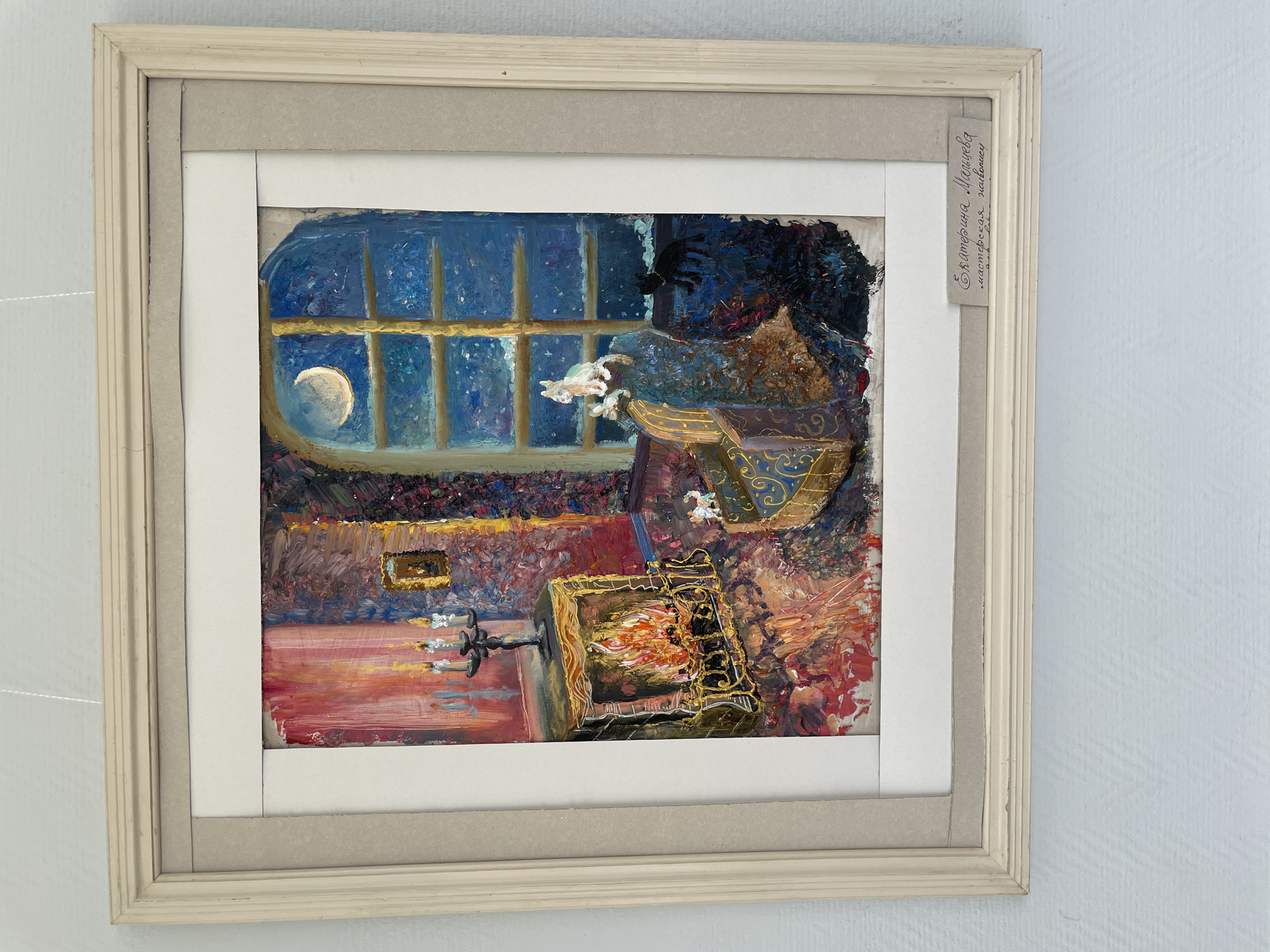 This is a framed painting depicting a cozy interior scene at night. A fireplace with burning logs is on the left, with three lit candles on a candelabra above it. In the center, a patterned armchair is placed near a large window. Two small white dogs are visible, one sitting on the armrest of the chair and another near the fireplace. A black cat is depicted outside the window on the floor. Through the window, a crescent moon is visible in a starry night sky, with snow or frost on the windowsill. The painting has a thick impasto texture. A small, handwritten label below the painting reads "Екатерина Мальцева, мастерская 'Полиглот'". This translates to "Ekaterina Maltseva, 'Polyglot' workshop". The image itself is taken from an angle, with a light blue, textured wall visible behind the framed artwork. The frame is made of light-colored wood with a simple, layered design. There are no discernible landmarks or specific architectural elements that would indicate a location in the city of Perm, Russia, beyond the artist's signature.
