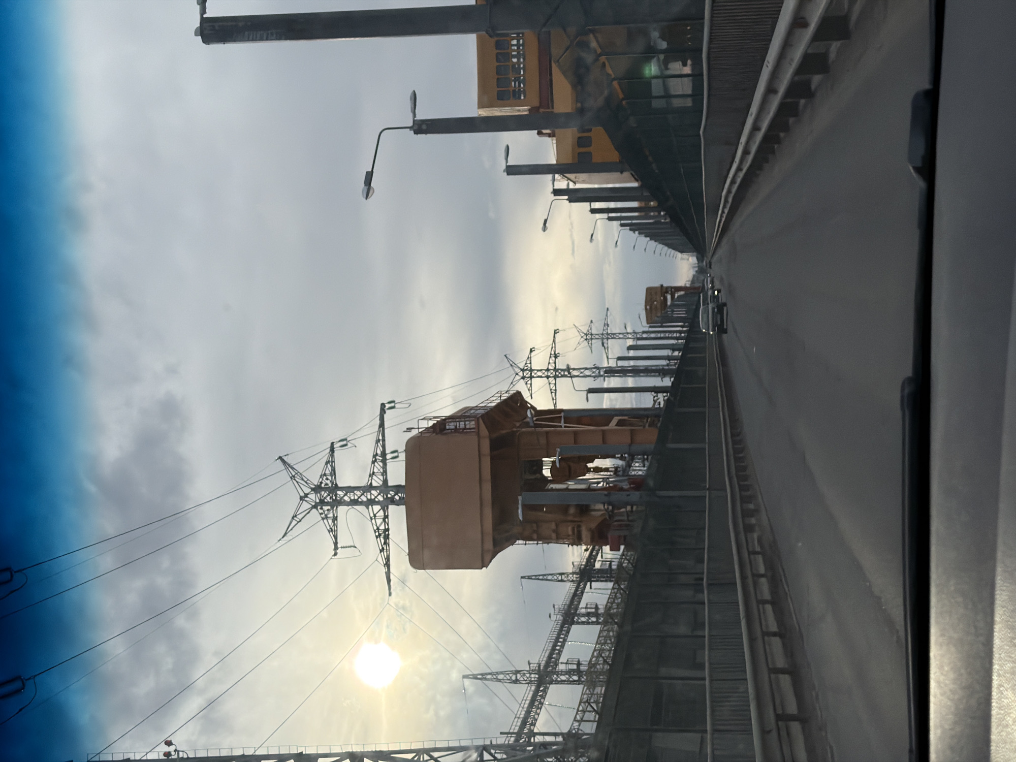The image, captured from a vehicle's interior looking forward, depicts a wide, elevated road or bridge extending into the distance. The view suggests driving along a significant industrial complex.
To the left of the road, a complex network of large electricity pylons and high-voltage power lines dominates the scene. Further left, a prominent, massive industrial structure, colored brown or orange, stands tall, flanked by other intricate metal constructions and smaller pylons. The bright, circular glare of the sun is visible low on the left, partially obscured by clouds, indicating daytime.
On the right side of the road, a concrete barrier or guardrail separates the roadway from a series of industrial buildings, some with distinct yellow facades, which line the structure. Streetlights on poles are visible along both sides of the road and within the industrial area. A single vehicle can be seen in the far distance on the road.
The sky is largely overcast with varied cloud formations, allowing sunlight to break through in places. The presence of numerous power lines, large industrial structures, and the road strongly suggests a substantial power generation or transmission facility. Given the context of Chaikovsky, Russia, the scene likely depicts the area around the Votkinsk Hydroelectric Power Station. No discernible text is visible.