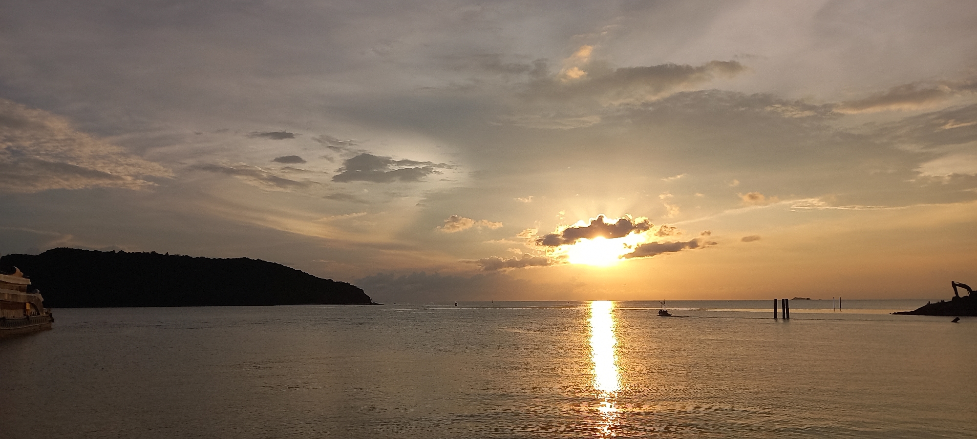 The image shows a sunset over the water in Thong Sala, Thailand. In the foreground is the calm sea reflecting the sunlight. A silhouette of an island is visible on the left. A small boat is making its way on the water. In the distant right, there appears to be a small construction site. The sky is cloudy with streaks of light shining through.