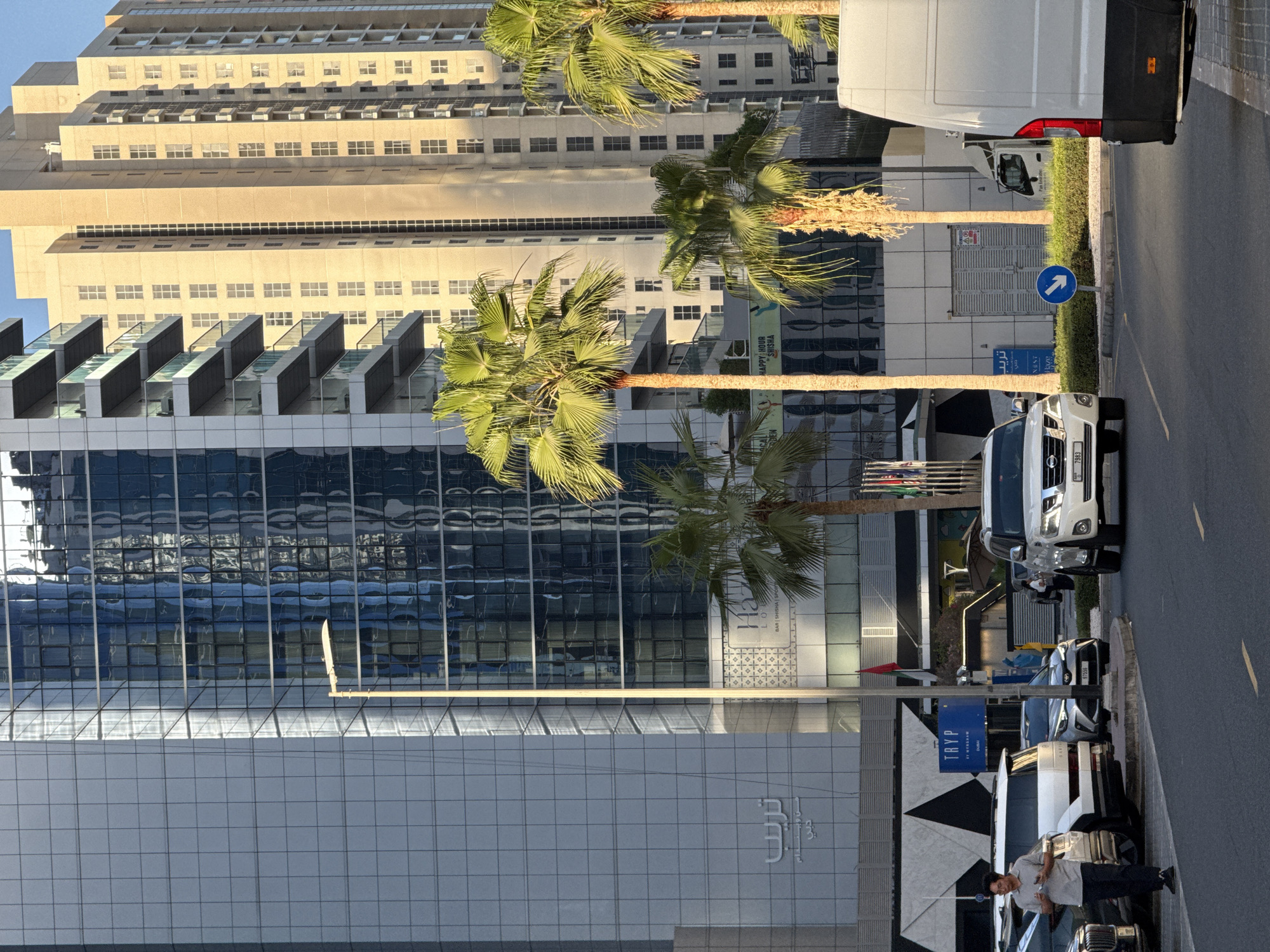 This is a street-level view in Dubai, United Arab Emirates, featuring modern architecture and palm trees. In the foreground, a dark asphalt road curves to the right, with yellow lane markings visible. On the left side of the road, several parked vehicles are present, including a white SUV, a silver sedan, and the rear of a white van on the far right. A man in a light-colored shirt and dark pants is walking away from the camera, holding a phone.
Behind the vehicles, a large building with a grid of reflective blue windows dominates the left side of the frame. To its right, a lighter-colored, taller building with many square windows stretches upwards, catching the sunlight. Several palm trees are interspersed between the buildings and in front of them, with lush green fronds and some showing signs of dryness.
A street lamp with a contemporary design stands near the blue-windowed building. Signage is visible on the lower levels of the buildings, including a blue sign that reads "TRYP BY WYNDHAM DUBAI." Another sign, partially obscured, appears to be in Arabic script. Flags are also visible, including a red flag on a pole. The overall impression is an urban environment characterized by contemporary design and the presence of tropical vegetation.