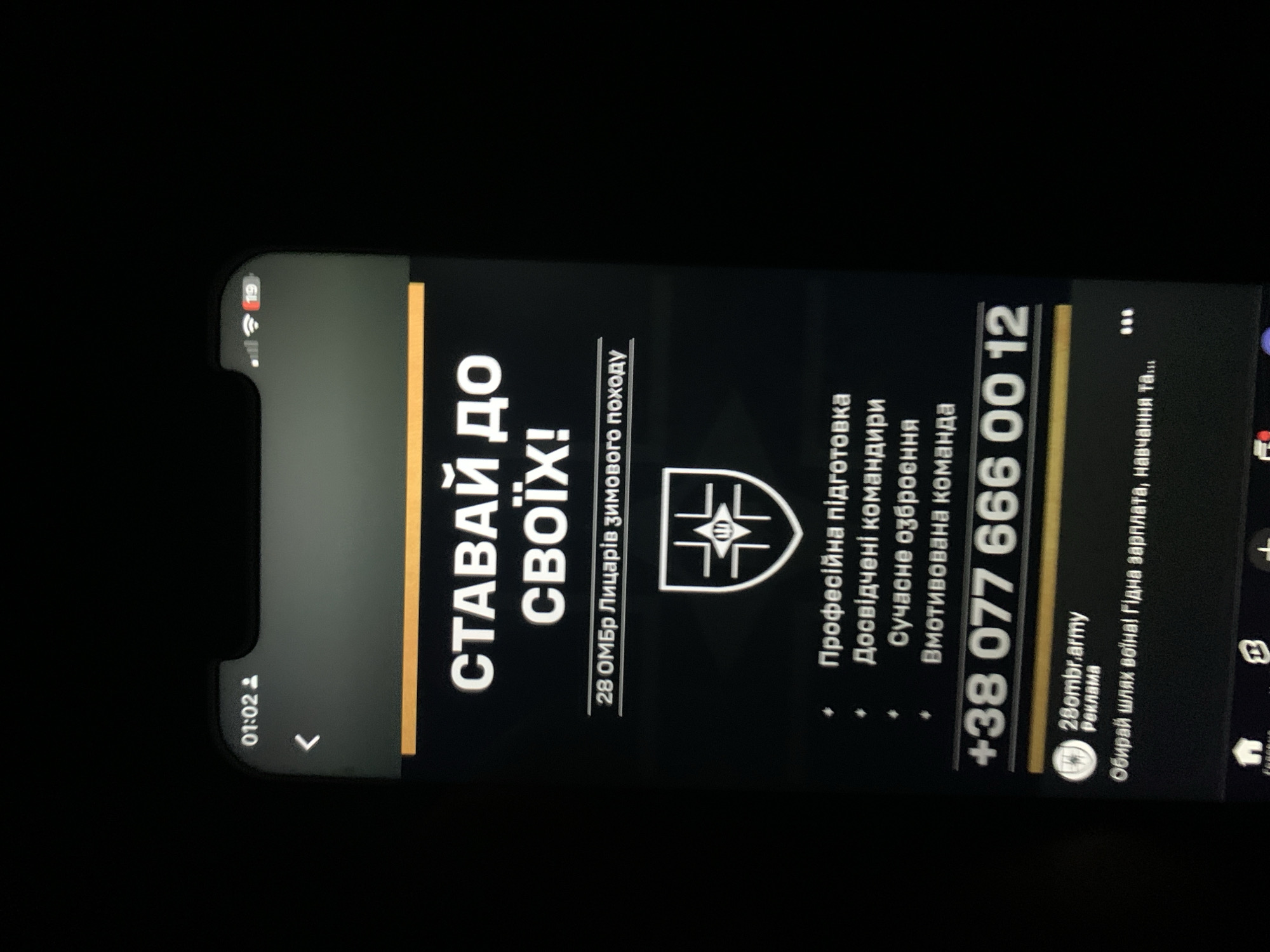 The image shows a close-up of a smartphone screen displaying an advertisement. The advertisement is in Ukrainian and features the text "СТАВАЙ ДО СВОЇХ!" which translates to "JOIN YOUR OWN!". Below this, it states "28 ОМБр Лицарів зимового походу" meaning "28th Mechanized Brigade Knights of the Winter Campaign". A shield emblem with a stylized symbol is shown, followed by bullet points listing "Професійна підготовка" (Professional training), "Досвідчені командири" (Experienced commanders), "Сучасне озброєння" (Modern weapons), and "Вмотивована команда" (Motivated team). A phone number, "+38 077 666 00 12", is prominently displayed. At the bottom, there is a profile picture with the text "28ombr.army Реклама" (Advertisement) and a tagline reading "Обирай шлях воїна! Гідна зарплата, навчання та..." (Choose the path of a warrior! Decent salary, training and...). The top of the phone shows the time "01:02" and signal indicators. The background of the image is dark, suggesting the photo was taken in low light conditions. There is no information in the image that can be used to determine its location in Zhmerynka, Ukraine.