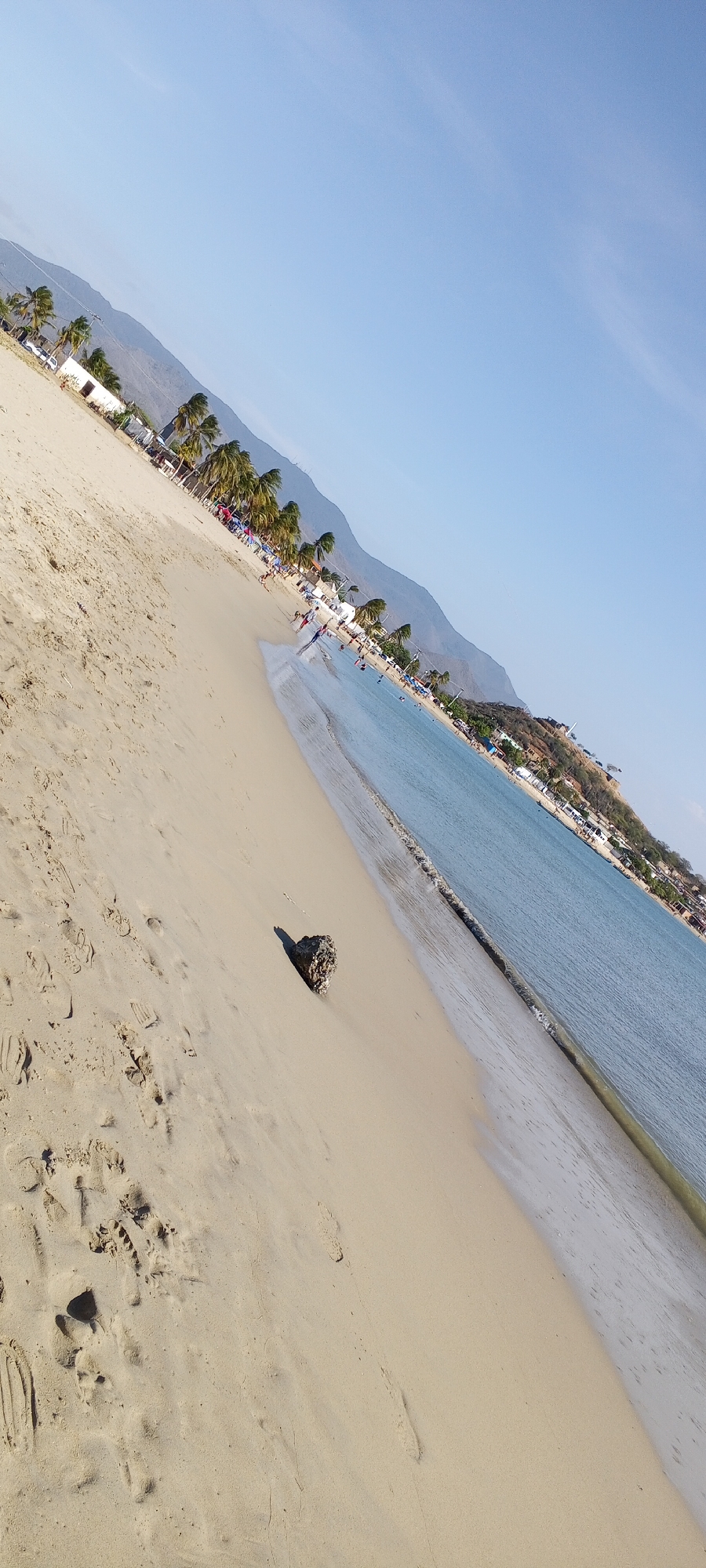 The image depicts a beach scene. A sandy beach occupies the foreground, with footprints visible in the sand. A dark-colored rock sits on the beach near the water's edge. The water is calm and light blue, with a subtle wave line along the shore. In the distance, a coastline is visible with buildings and vegetation. Further inland, mountains rise against a clear blue sky. Along the beach are palm trees, people, and tents.