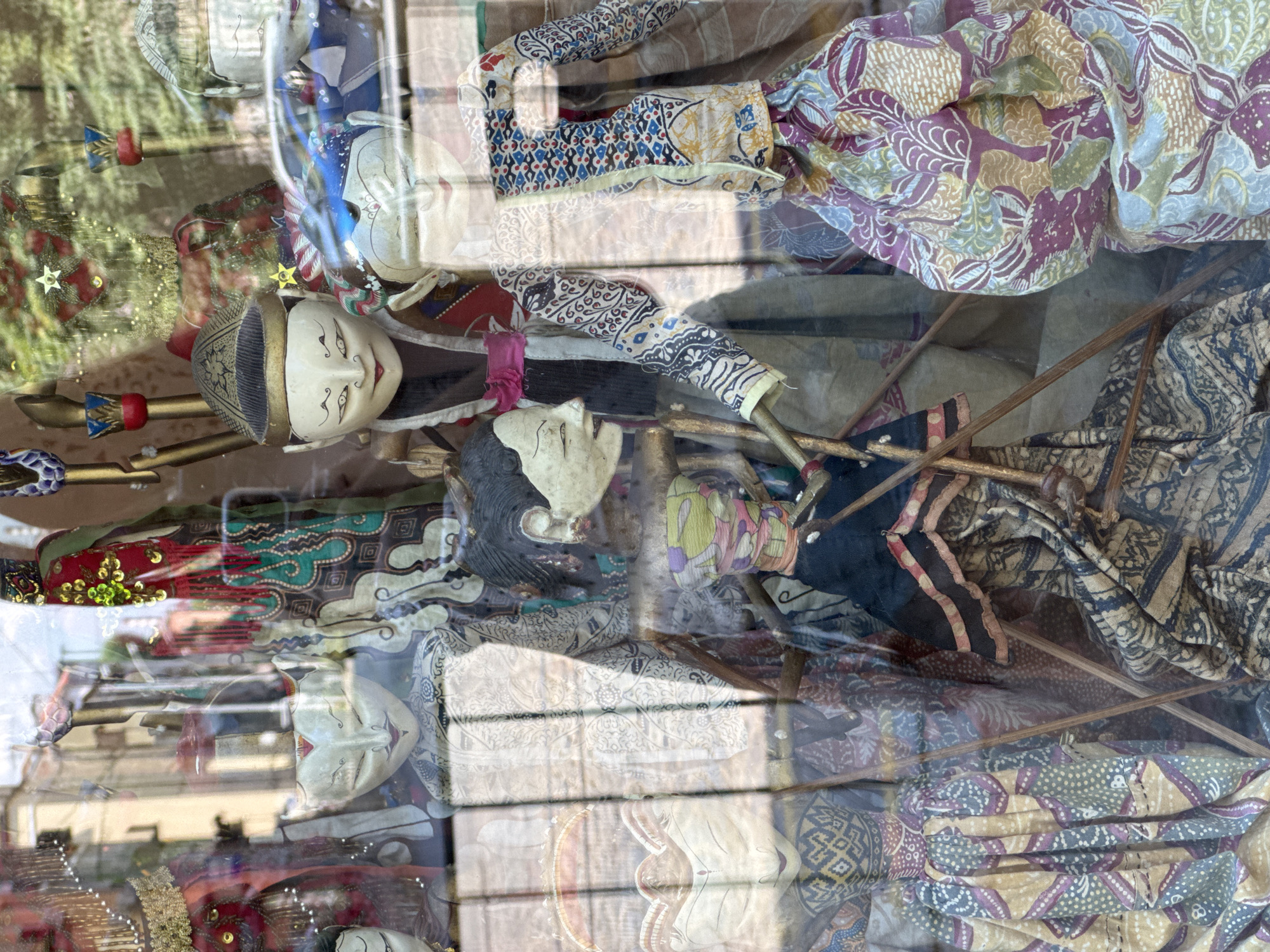 The image shows a display of traditional Indonesian wayang kulit puppets in a shop window in Sofia, Bulgaria. The puppets, made of carved leather and fabric, are adorned with intricate patterns and vibrant colors. They are arranged in a theatrical setting, with wooden rods used to manipulate their movements. The window reflects the street outside, with glimpses of buildings and a car, indicating an urban environment. The puppets are the primary focus, with their detailed masks and elaborate costumes visible through the glass. The lighting suggests it is daytime.