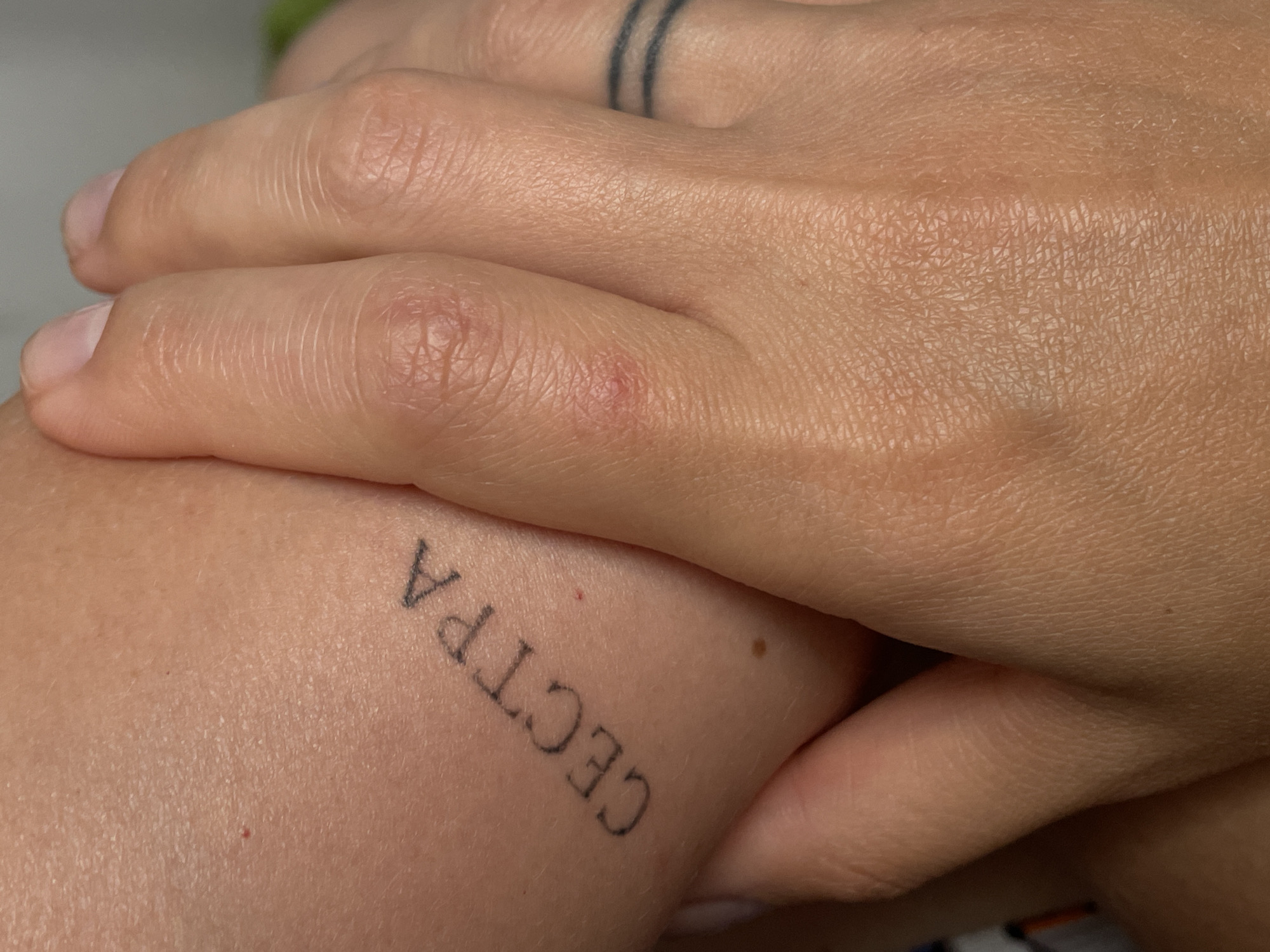 A close-up shot focuses on a person's arm and hand. On the arm, the word "СЕСТРА" (sister in Russian) is tattooed in black, block letters. The skin appears lightly tanned with visible pores and texture. A small red mark, possibly a healed scratch or mole, is present near the tattoo. The hand is positioned over the arm, obscuring part of the tattoo and skin. The fingers are slightly curled, and the skin on the fingers shows natural creases and texture. A black, double-ring tattoo is visible on one of the fingers. The background is blurred and appears to be an indoor setting with neutral tones. The image does not contain any elements that indicate a specific location in Rostov-on-Don, Russia.