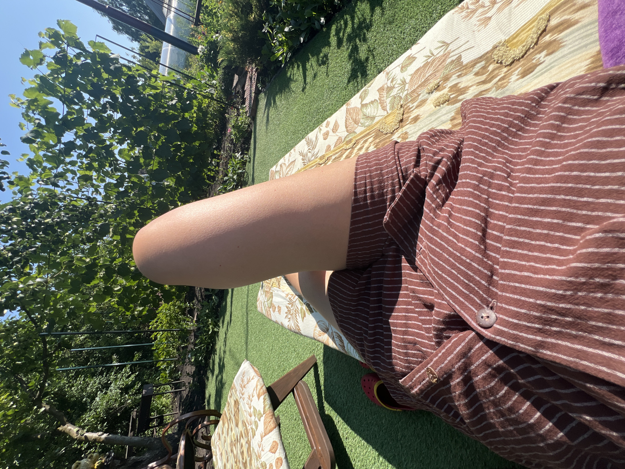 A person lies on a floral-patterned lounge chair on a patch of artificial turf. Their legs are extended, with the right leg prominently featured, and they are wearing a brown and white pinstripe garment. Red clogs are visible on the left foot. To the left of the lounge chair, a similar floral-patterned table is positioned. The background shows a lush green garden with trees and foliage, suggesting an outdoor setting. The scene takes place in Lopatino, Russia, on a sunny day.