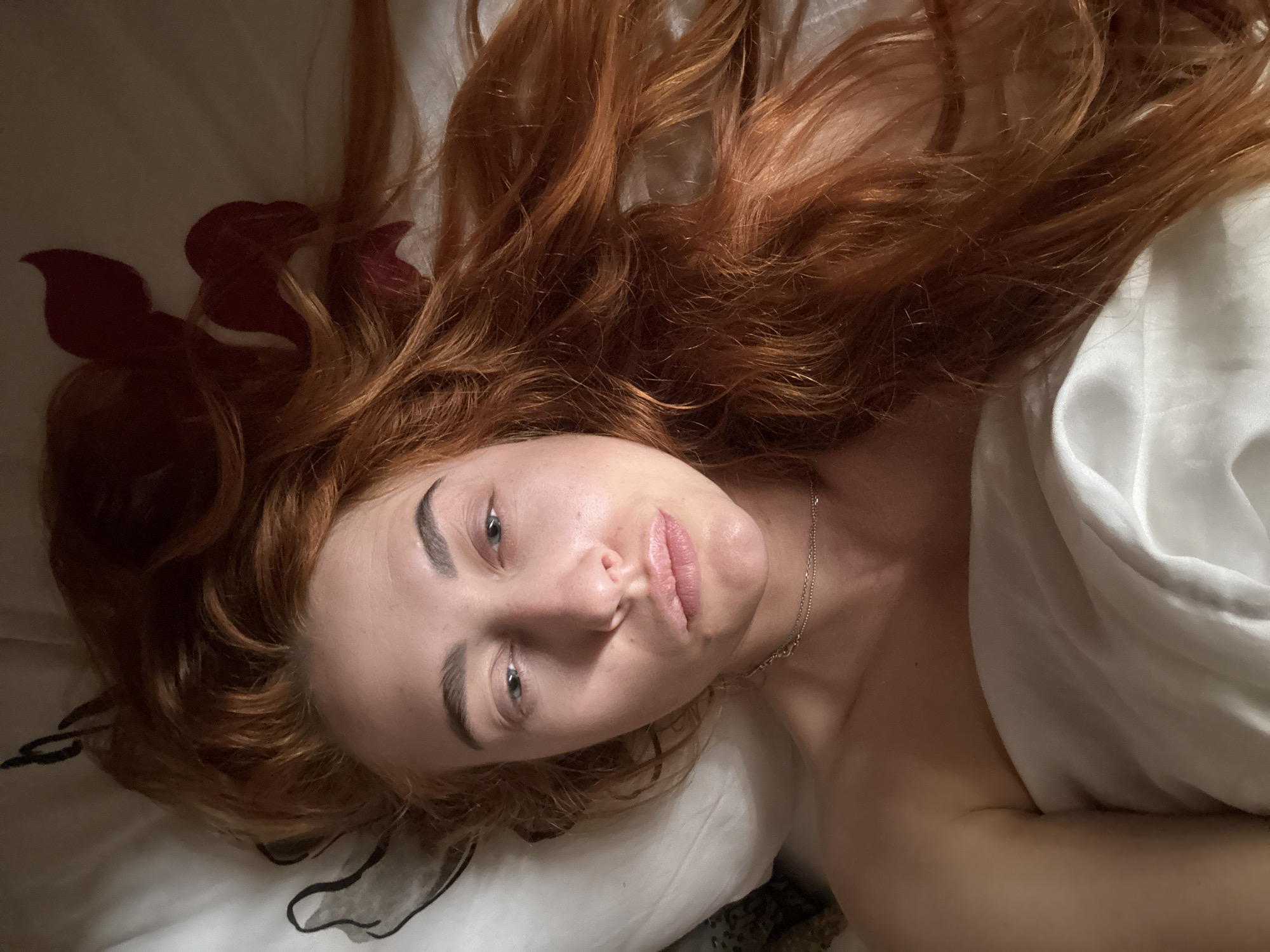 A light-skinned person with long red hair is lying on a white pillow with a white sheet partially covering their body. A delicate silver chain necklace is visible around their neck. The image appears to be taken from a low angle, looking down on the person's face. There is no visible indication of the location being Budva, Montenegro. A red design can be seen in the lower left corner of the image.