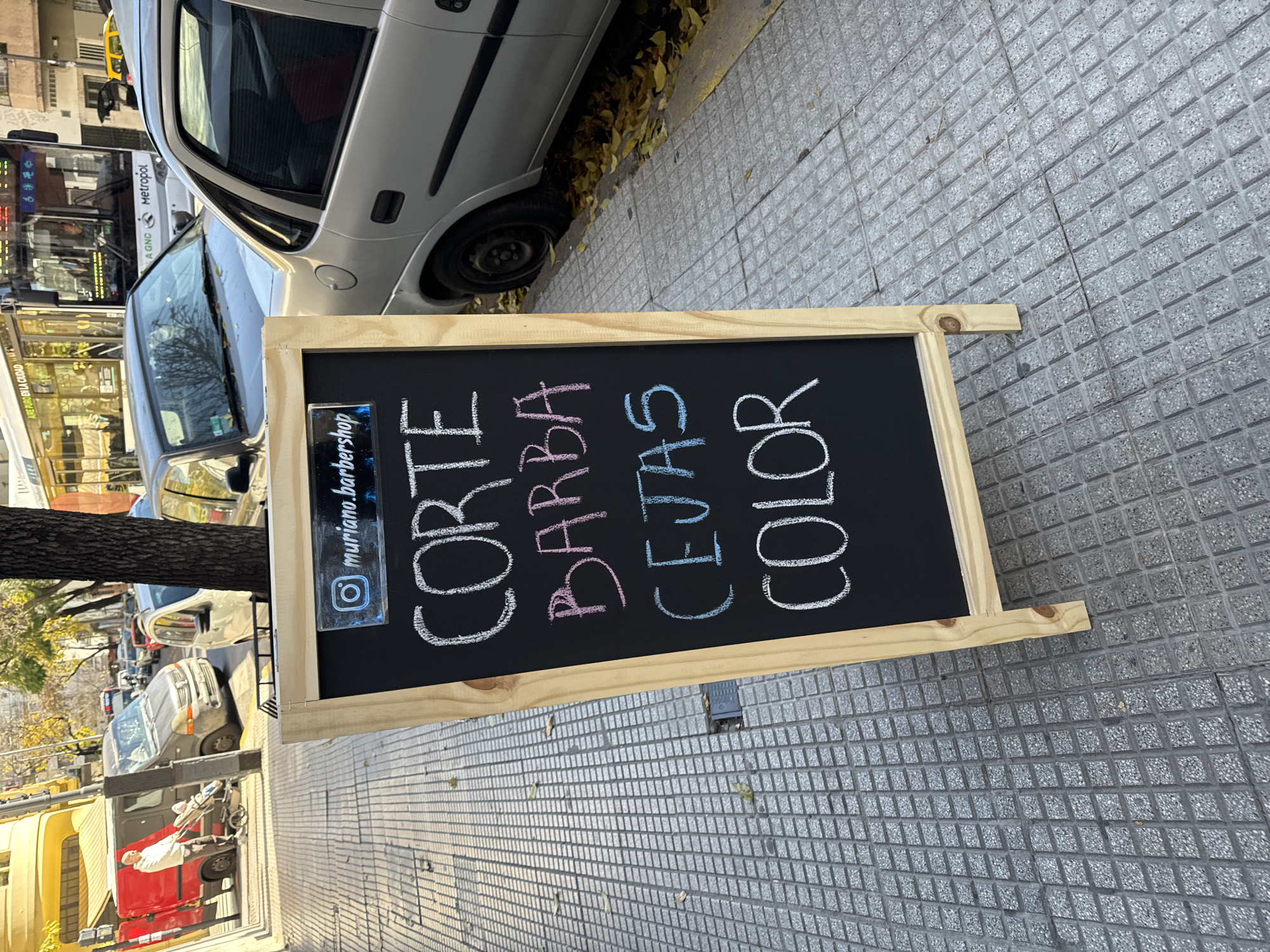 A street scene in Buenos Aires, Argentina, depicts a sidewalk with a gray textured tiled surface. A silver-colored car is partially visible on the left side of the frame, parked at an angle. A wooden-framed chalkboard sign stands on the sidewalk, displaying the words "CORTE," "BARBA," "CEJAS," and "COLOR" in white, pink, and blue chalk lettering. A sticker with an Instagram logo and the handle "muriano.barbershop" is affixed to the board. In the background, other cars, buses, and buildings are visible. A tree trunk is visible in the middle ground of the shot.