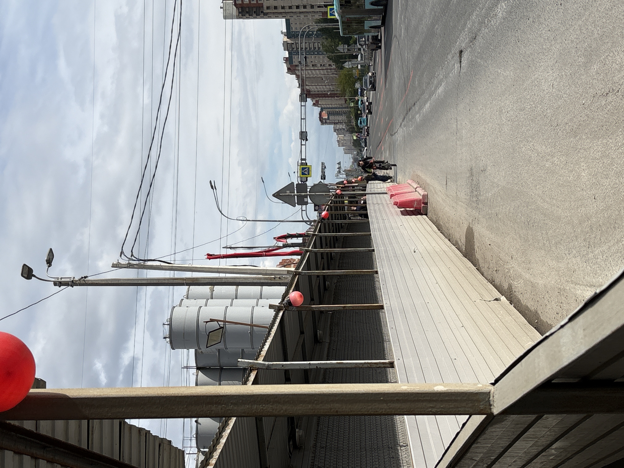 This image depicts an urban street scene in Saint Petersburg, Russia, under an overcast daytime sky. The perspective is from a slightly elevated position, looking along a wide asphalt road that recedes into the distance, flanked by multi-story buildings.
In the foreground on the left, there is an industrial or construction site. Several large, grey, cylindrical silos or storage tanks are prominent, rising above a structure with a corrugated metal roof and exposed support beams. A bright red spherical object is positioned on a beam in the immediate foreground, and red flexible hoses or pipes extend upwards from this area. Overhead, numerous power lines crisscross the sky, connected to utility poles, with streetlights attached to them.
The right side of the image is dominated by the asphalt road, which shows some faded white lane markings and newer red lines. Along the roadside, a line of red plastic barriers defines an area where several individuals are present. One person is seen riding a bicycle, while others stand or walk nearby. Further down the road, various vehicles, including cars and a bus, are visible, indicating active traffic.
Traffic infrastructure includes traffic lights suspended over the road and several road signs. One yellow diamond-shaped sign, often denoting a main or priority road, is clearly visible. Another blue square sign with a white figure indicates a pedestrian crossing. The scene conveys a typical urban environment with elements of construction or industrial activity alongside a busy thoroughfare.