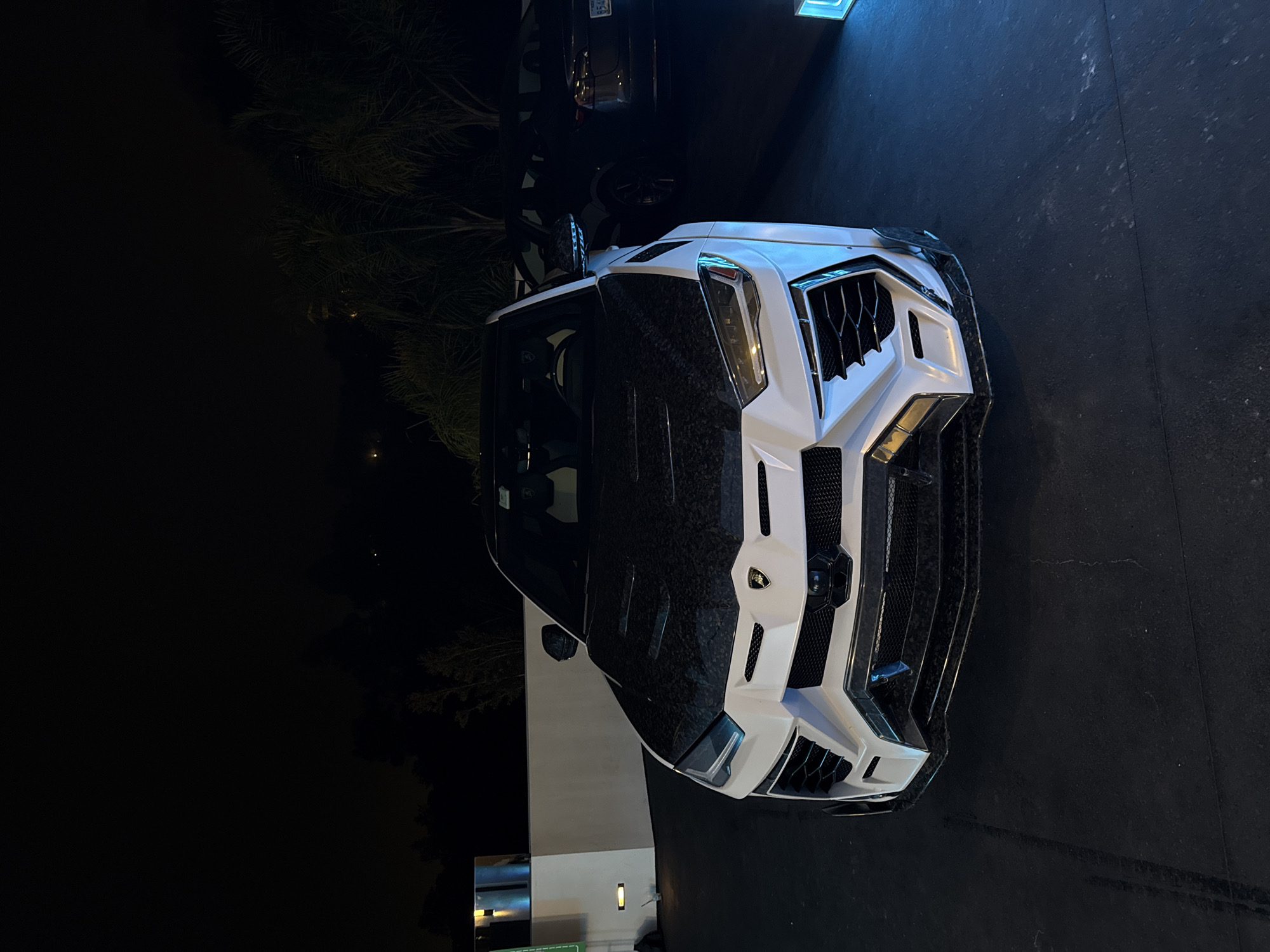 The image depicts a white Lamborghini Urus Mansory parked in a driveway. The front of the car is facing the camera, showcasing its distinctive aftermarket body kit. The hood and roof appear to have a carbon fiber finish. Another vehicle is partially visible behind the Urus, parked further back in the driveway. The environment suggests a residential area, with palm trees visible in the background and a building facade on the left side. Given the visible palm trees and architectural style, the location is consistent with a wealthy suburb within the city of Los Angeles, United States of America. The scene is illuminated by artificial lighting, possibly from street lamps or the building's exterior lights, as it appears to be night. There are no visible people in the image.