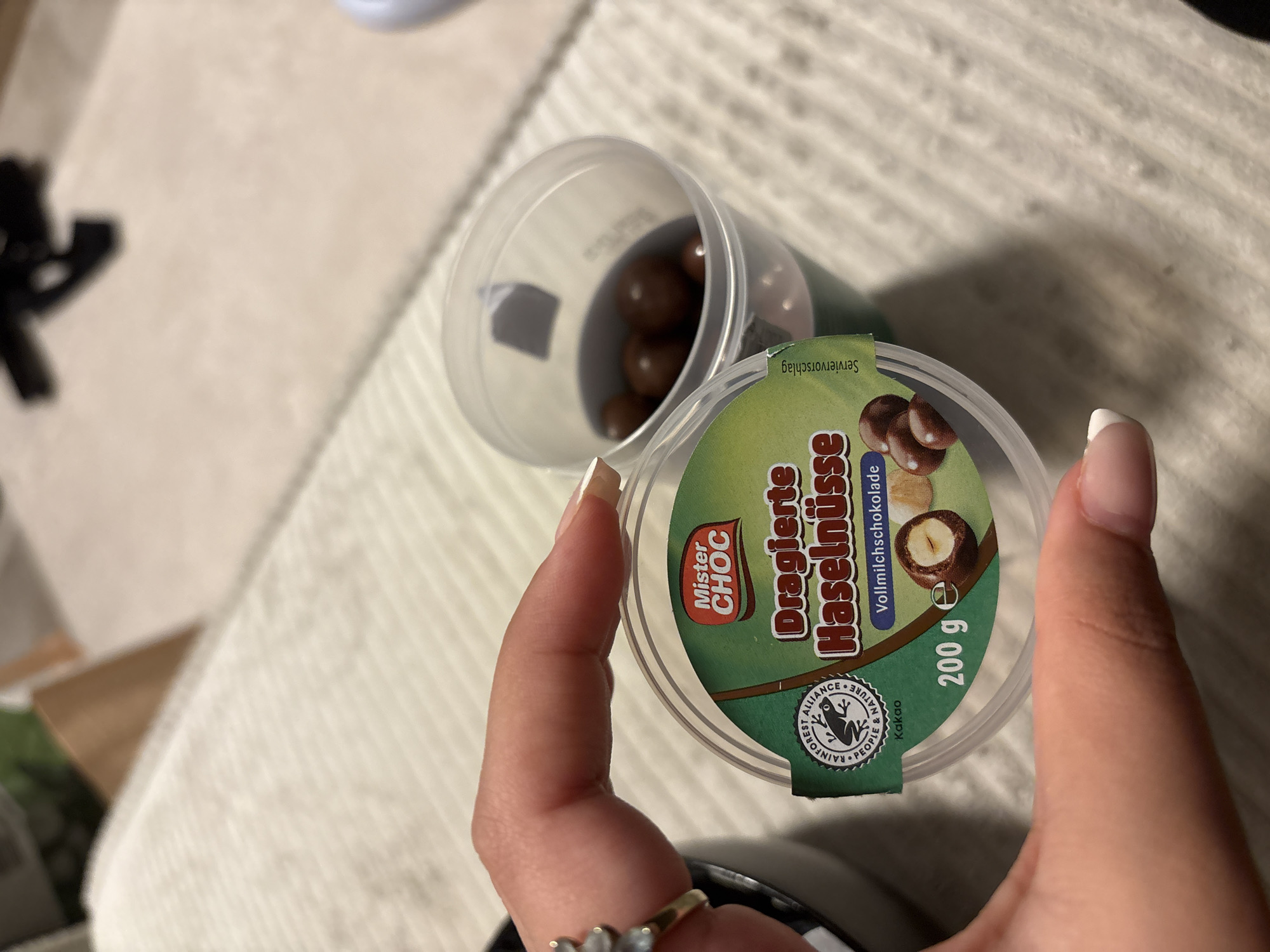 The image shows a partially open container of "Dragierte Haselnüsse" (chocolate-covered hazelnuts) produced by the brand "Mister Choc." The container is a clear plastic cup with a green and white label on the lid. The label includes the product name, an image of hazelnuts coated in chocolate, the brand logo, a Rainforest Alliance seal, and the weight, 200 grams. A hand holds the lid, partially revealing the chocolate-covered hazelnuts inside the container. The background features a textured, light-colored surface. The location of Mönchengladbach, Germany is not evident in the image, so the description cannot confirm nor deny such location.