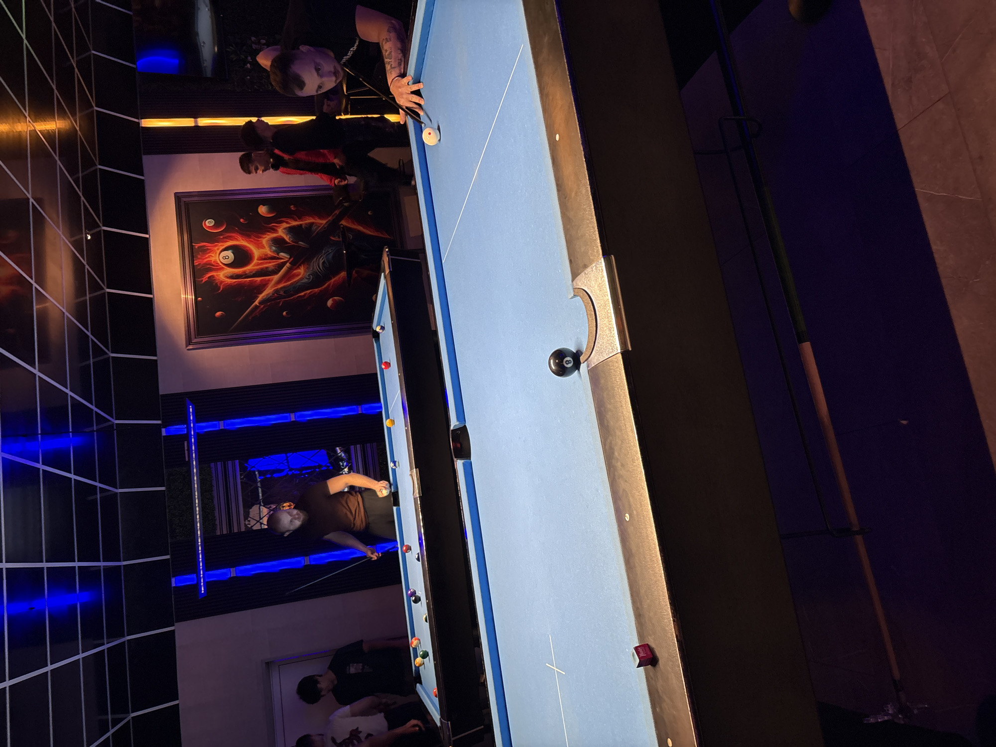 A well-lit interior features two pool tables, with one table prominently displayed with a white cue ball near the top, and a black 8-ball near the side pocket. Two men are seen leaning over this table, appearing to be playing. In the background, a mural adorns the wall. The second pool table is partially visible and features an ongoing game with multiple players. Blue lights illuminate a section of the background, suggesting a modern or entertainment venue. Given the prompt, the location is Ruse, Bulgaria.
